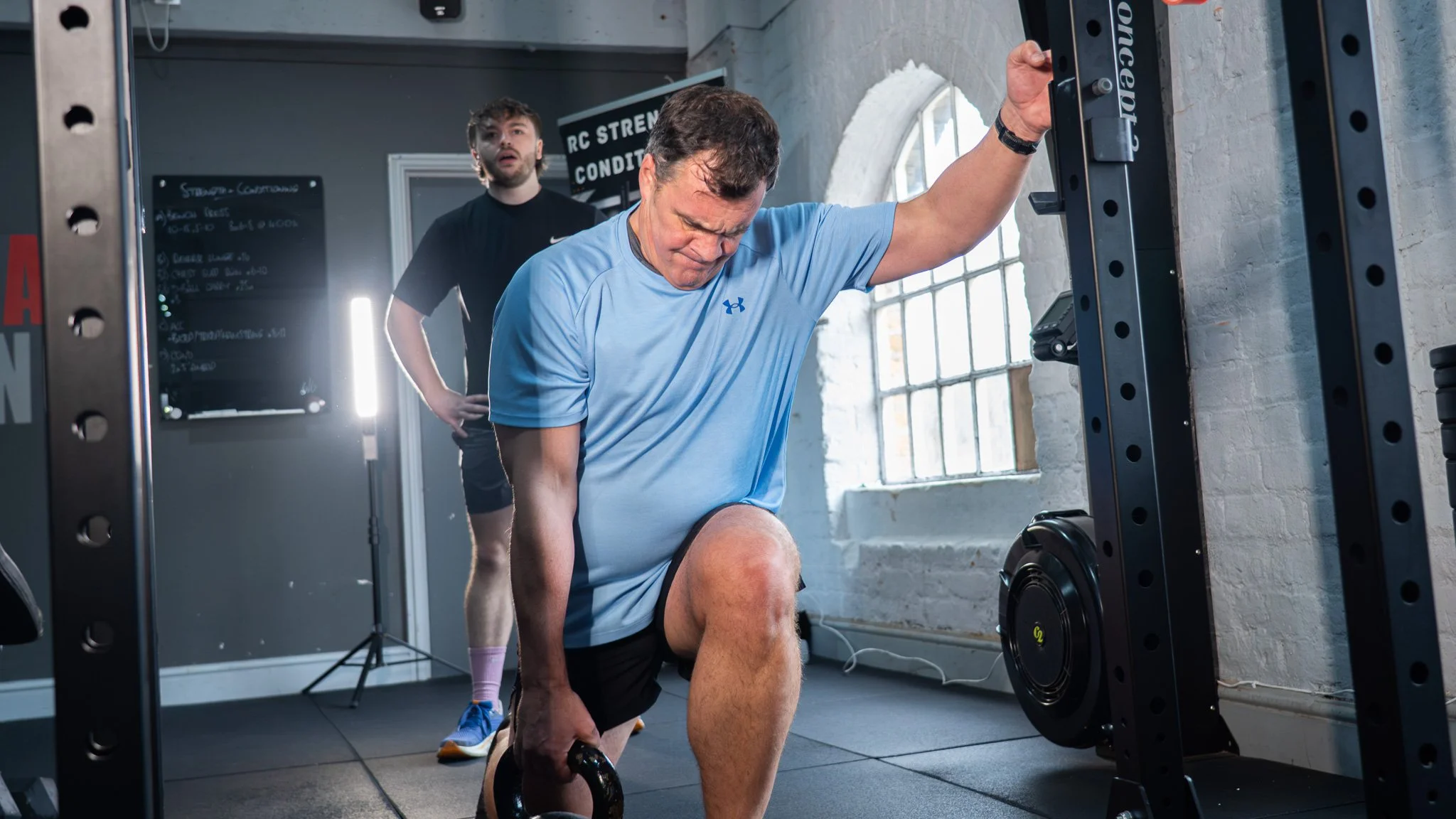 A man in a light blue athletic shirt and black shorts is performing a weighted lunge while holding a kettlebell in one hand in a gym. A spotter stands behind him, observing his form. The gym has large windows, white brick walls, and workout equipment.