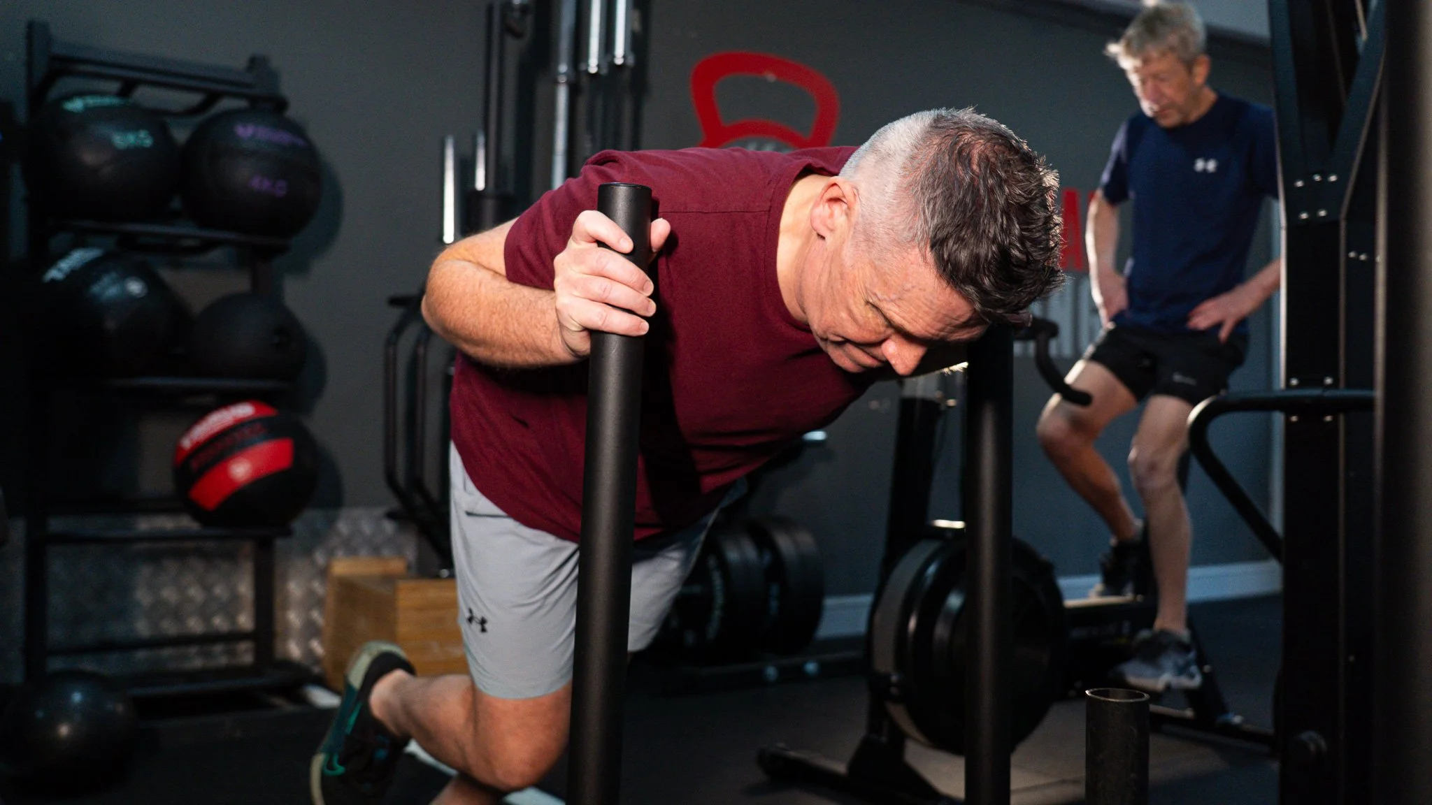 An elderly man with gray hair is doing a push-up exercise using a black hand gripper at a gym, with a trainer or fellow exerciser in the background.