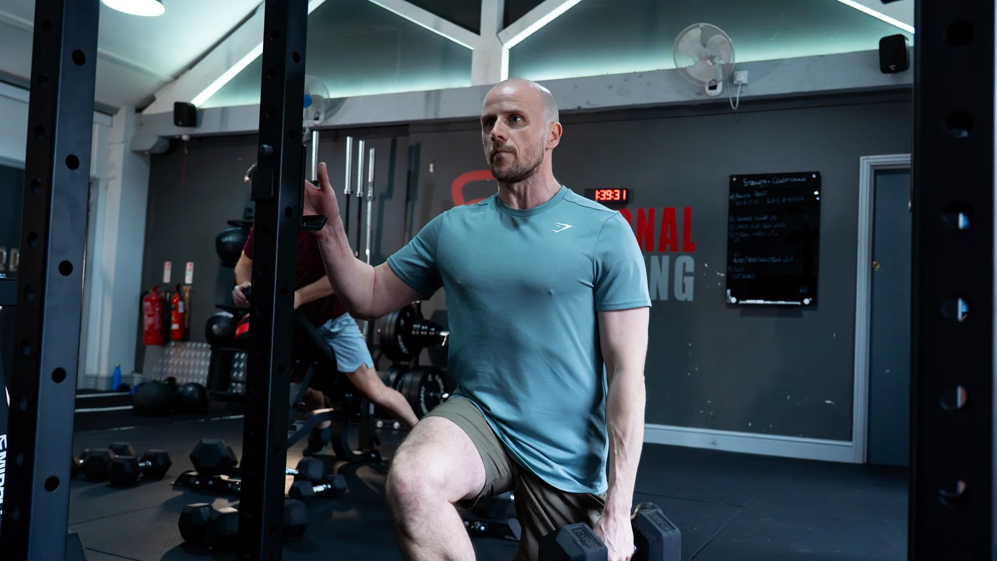 A man wearing a light blue athletic shirt and tan shorts kneeling with one knee on a workout bench in a gym, holding a dumbbell in each hand. He is surrounded by gym equipment, including weights and fire extinguishers, and appears focused during his workout.