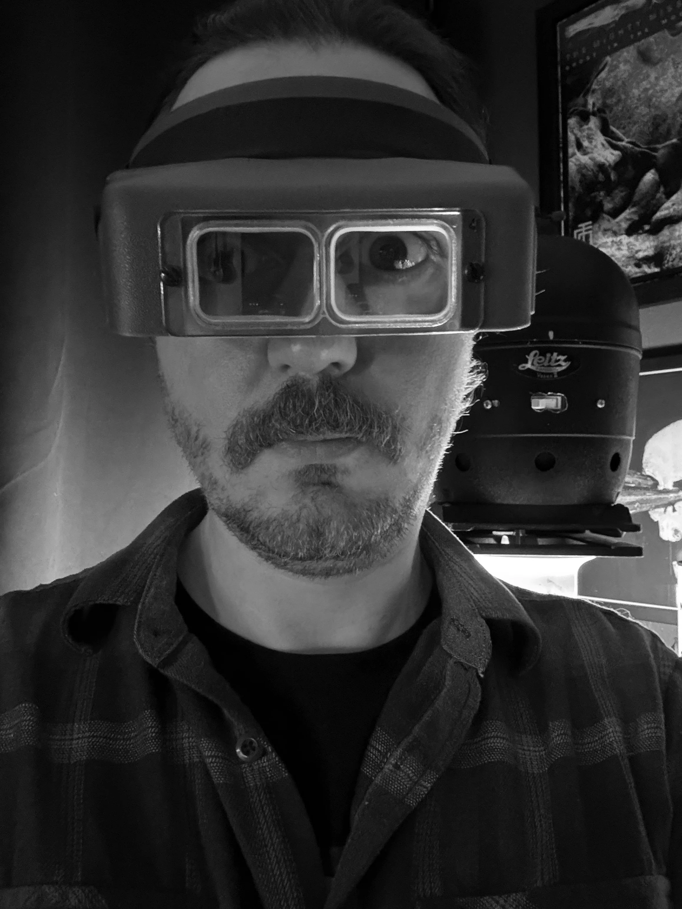 A man with a beard and mustache wearing protective goggles takes a selfie in black and white, with a dark background.