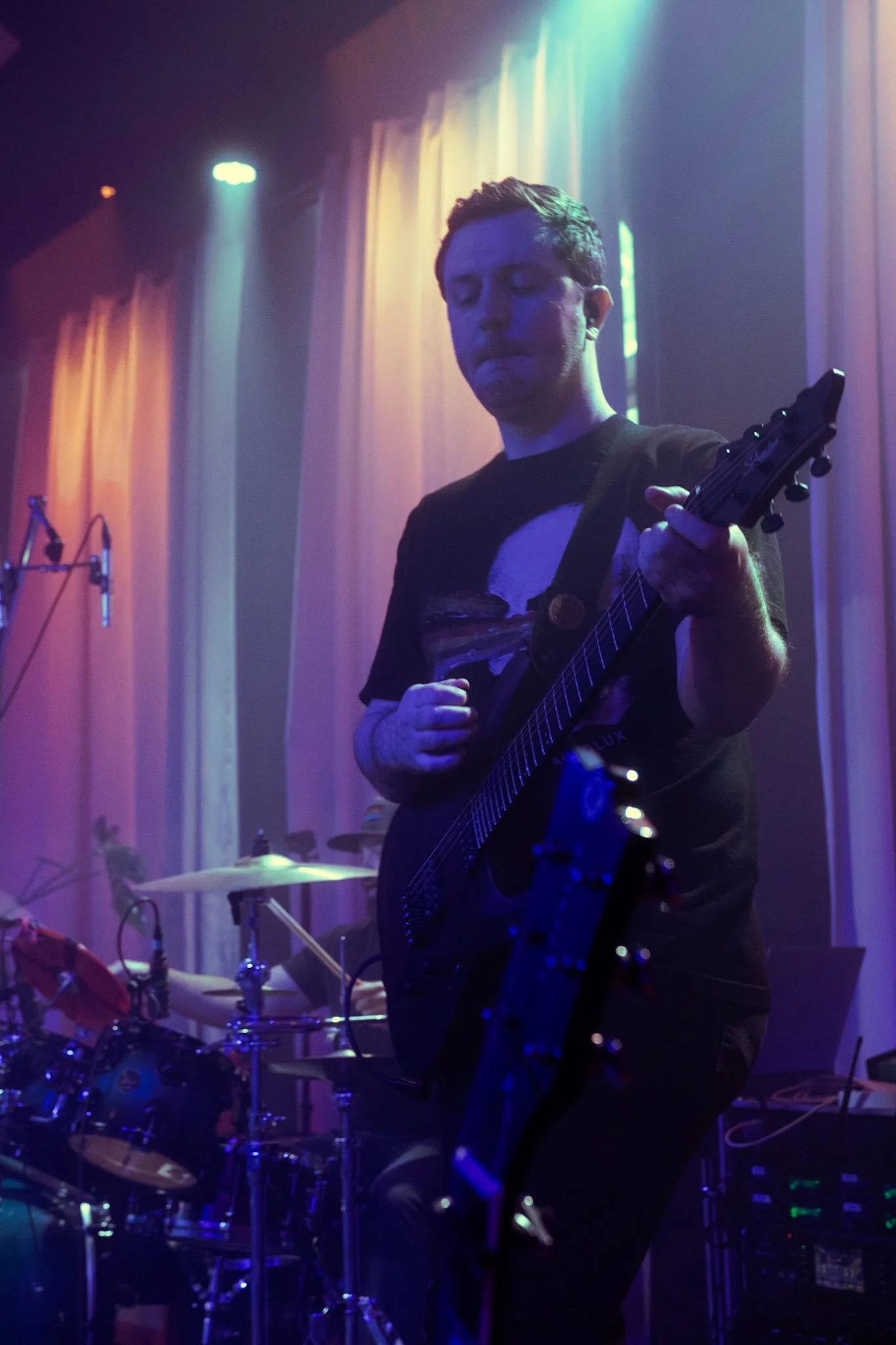A male guitarist performing on stage illuminated by colorful stage lights with curtains in the background.