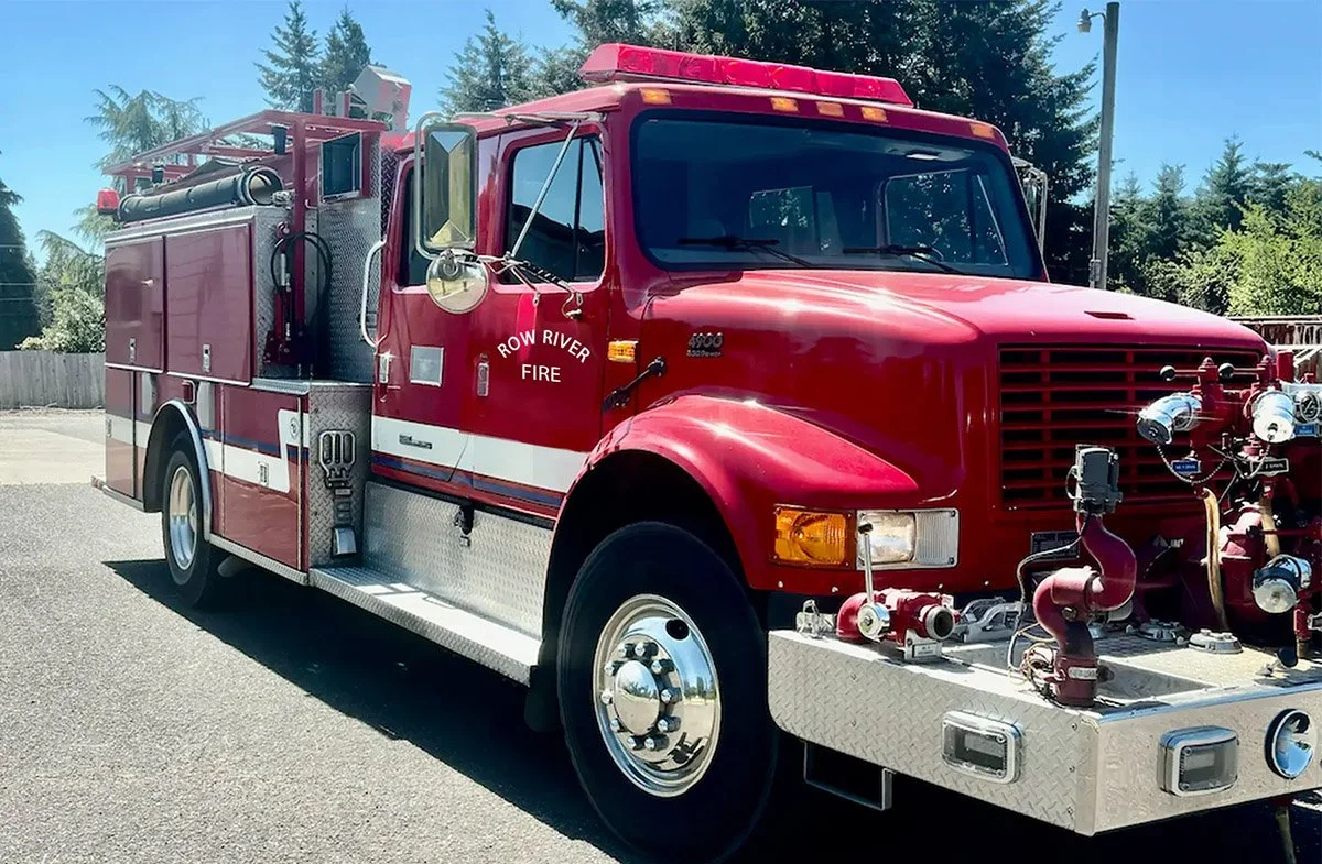 Red fire truck with white and blue stripes, labeled Row River Fire, parked outdoors under a clear blue sky.
