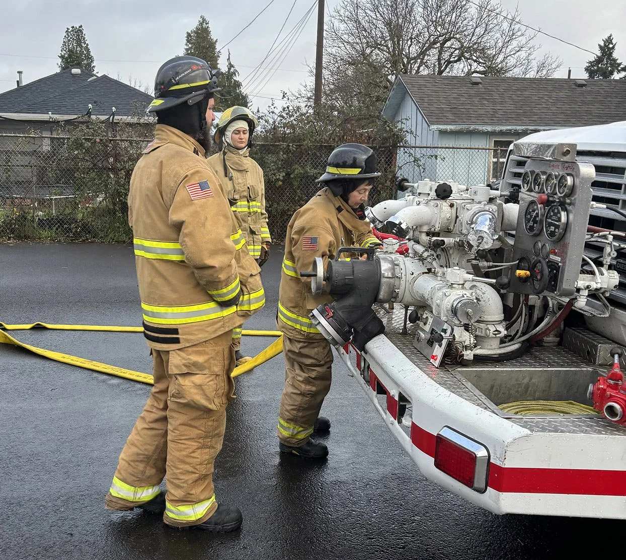 Three firefighters in tan uniforms and black helmets operate a fire truck's equipment on a wet street, with houses and trees in the background.