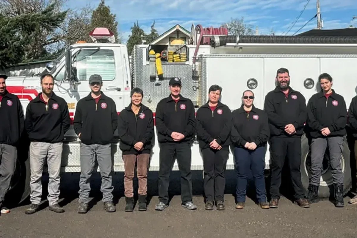 A group of nine firefighters standing in front of a fire truck on a sunny day.