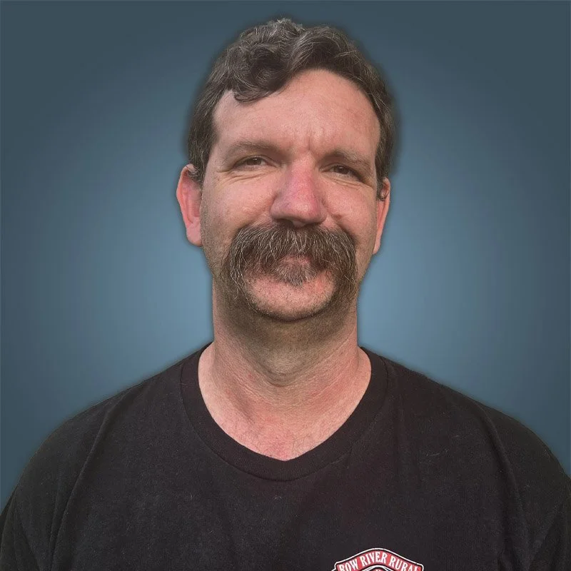 A middle-aged man with a mustache and short curly hair smiling at the camera, wearing a black T-shirt against a solid blue background.