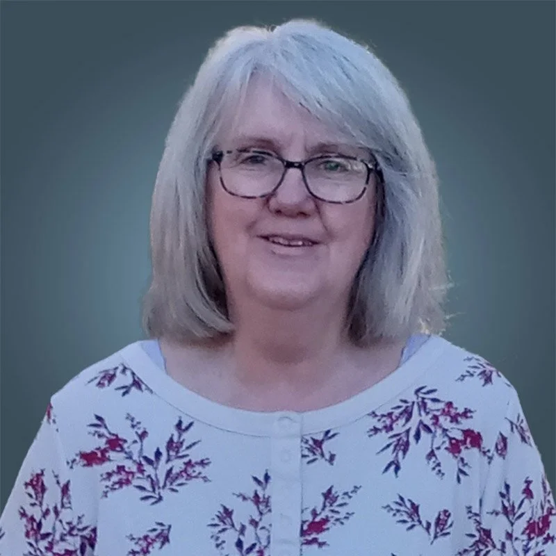 A middle-aged woman with shoulder-length gray hair and glasses, wearing a white shirt with a red and purple floral pattern, smiling outdoors against a blurred green background.