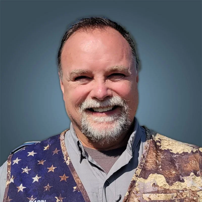 A smiling middle-aged man with a gray beard and short hair, wearing a shirt with a patriotic vest featuring the American flag design.