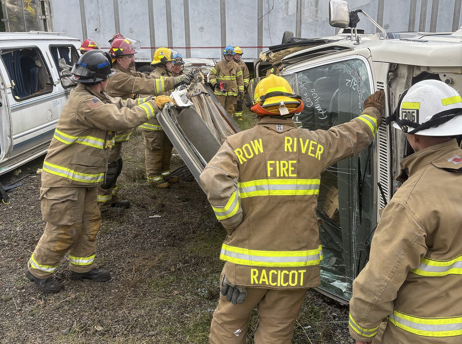 Firefighters from Row River Fire rescue a truck that has flipped onto its side in an accident scene. The firefighters are wearing yellow helmets and tan turnout gear with reflective stripes, working together to possibly free someone or stabilize the vehicle.