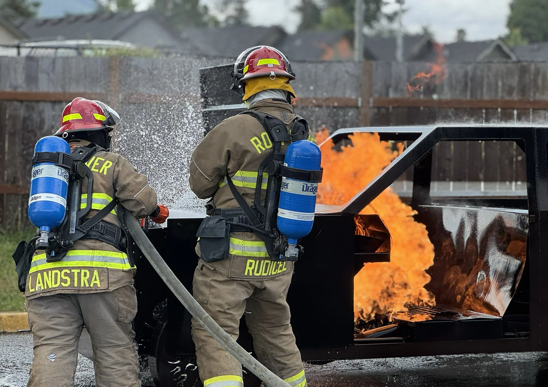 Two firefighters wearing protective gear and helmets, working to extinguish a car fire with flames visible inside the vehicle, on a residential street.