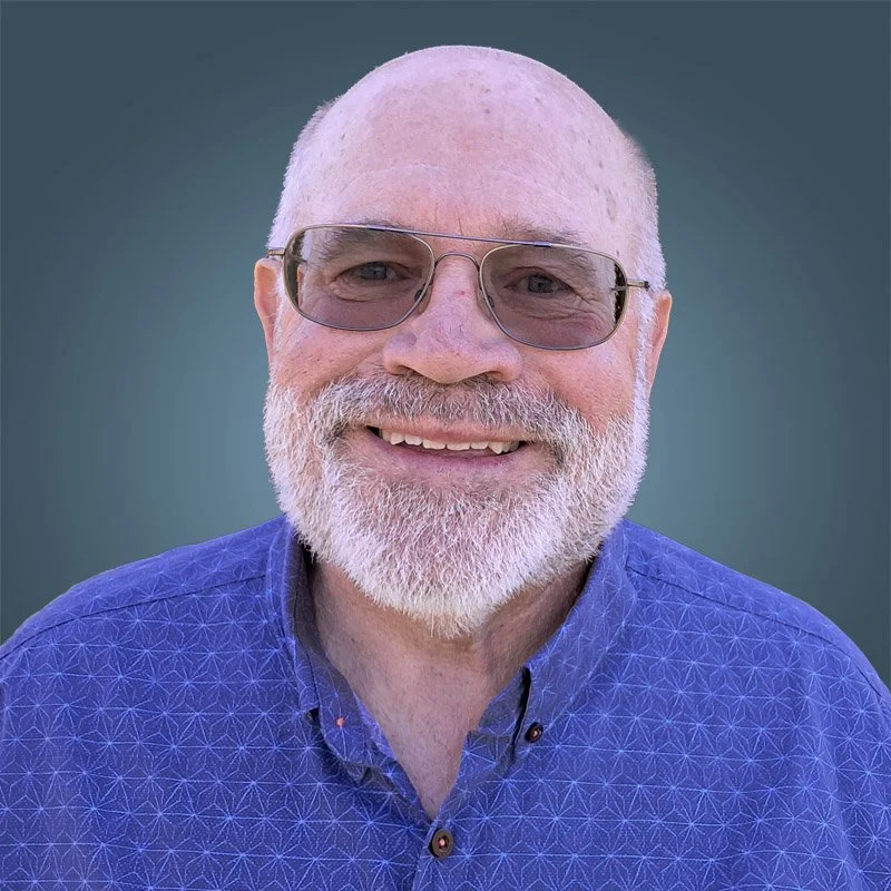 A smiling elderly man with a white beard and glasses, wearing a blue patterned shirt, standing against a plain background.