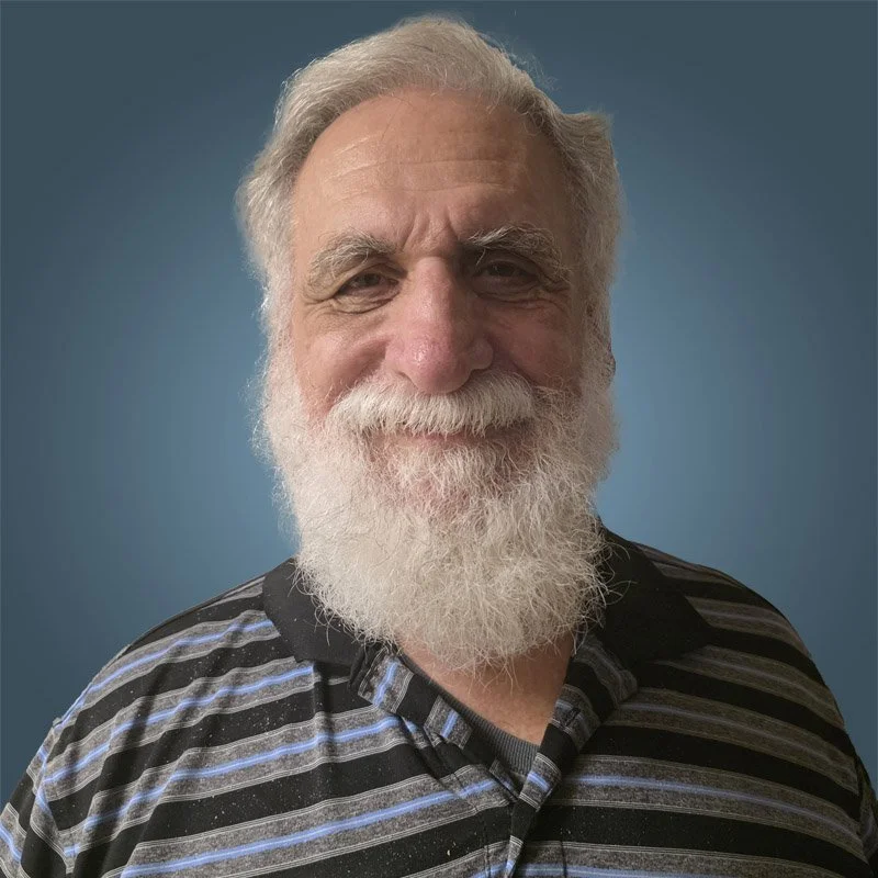 A senior man with a full white beard and hair, smiling, wearing a black and gray striped collared shirt against a blue background.