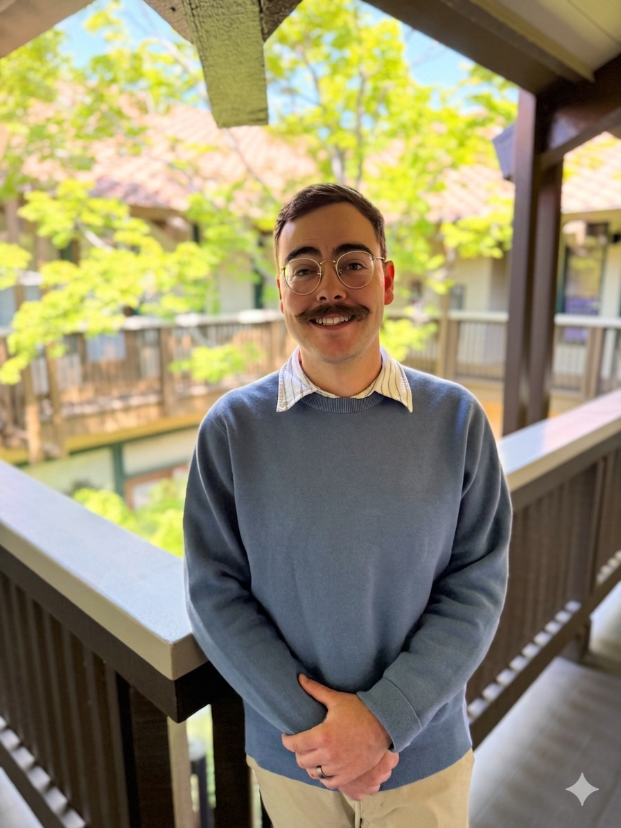 A man with glasses and a mustache smiling, standing on a balcony with green trees and houses in the background.