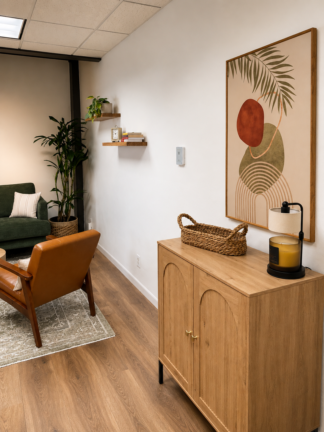 Living room corner with a wooden sideboard, decorative basket, abstract wall art, lamp, a tall plant, and seating area with a green sofa and a leather armchair.