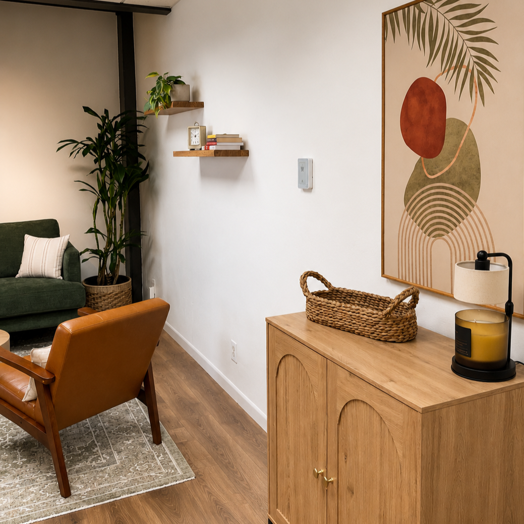 green sofa, an orange armchair, and a patterned area rug. There are wooden shelves with books and a plant on the wall, a wicker basket on a wooden cabinet, and abstract artwork hanging nearby. A lamp and a candle are on the cabinet, and there's a large potted plant near the sofa.