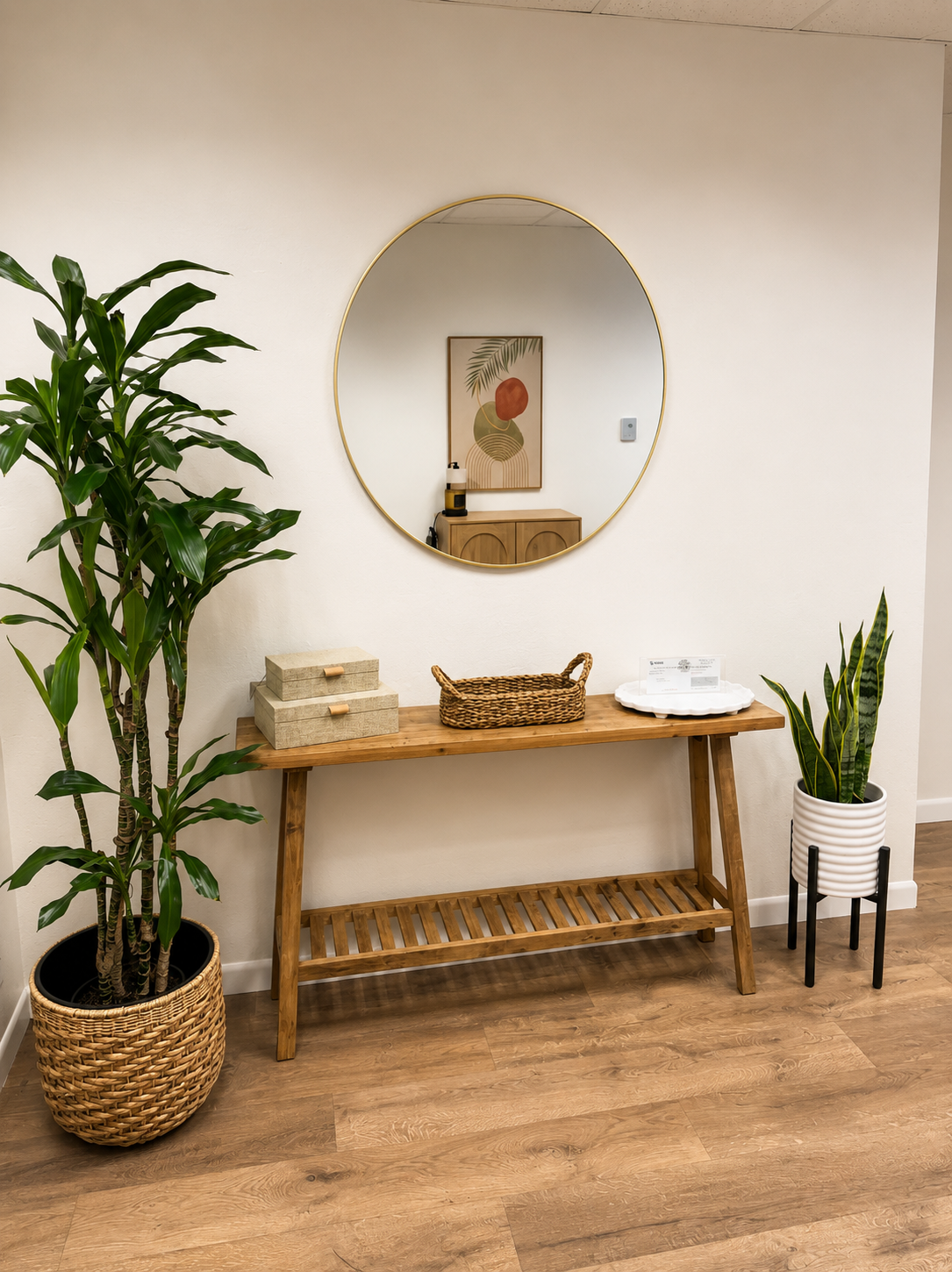 Interior scene with a wooden console table, a large round mirror, green potted plants, decorative boxes, and a framed artwork on the wall.