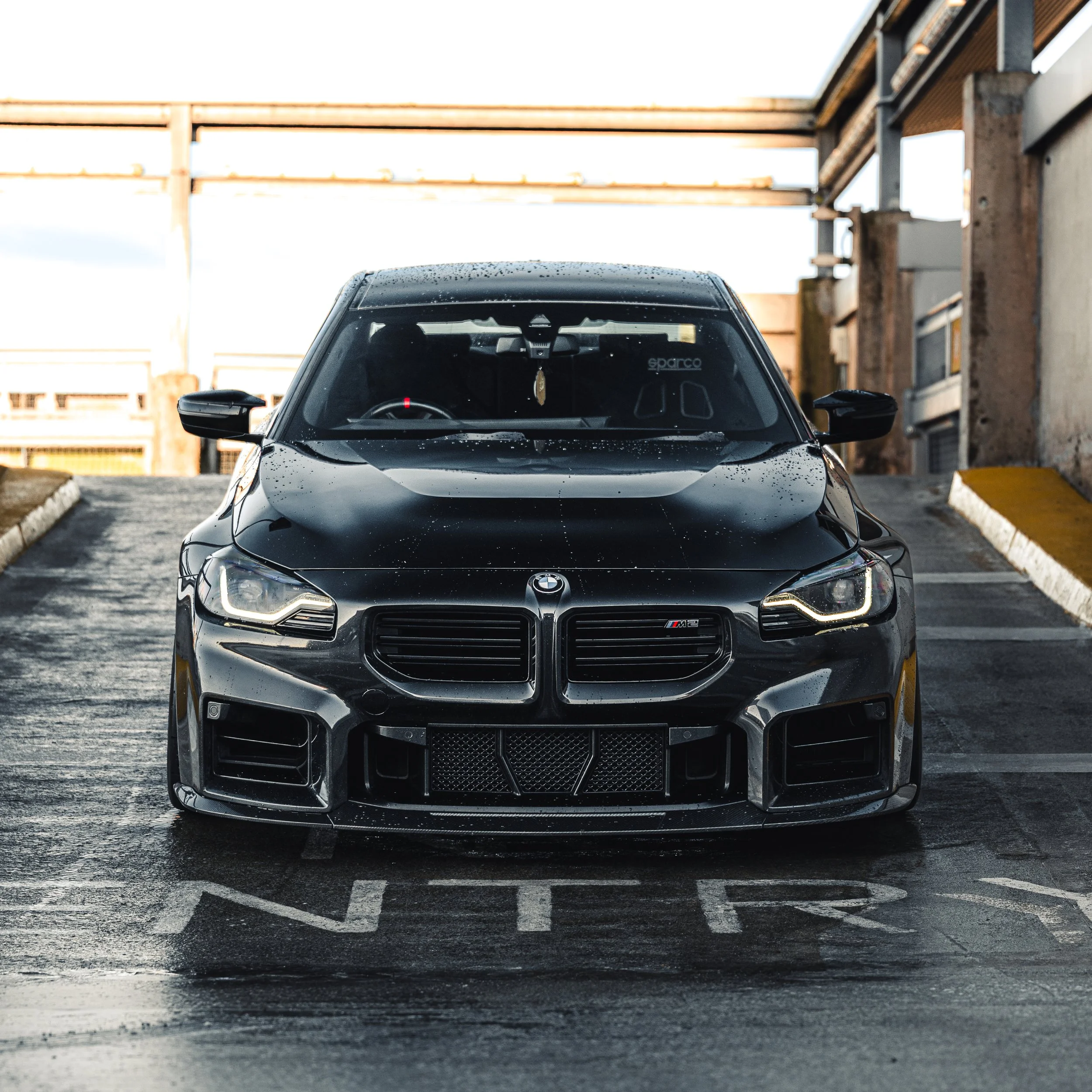 A black BMW M2 parked in an outdoor parking lot with industrial buildings in the background.