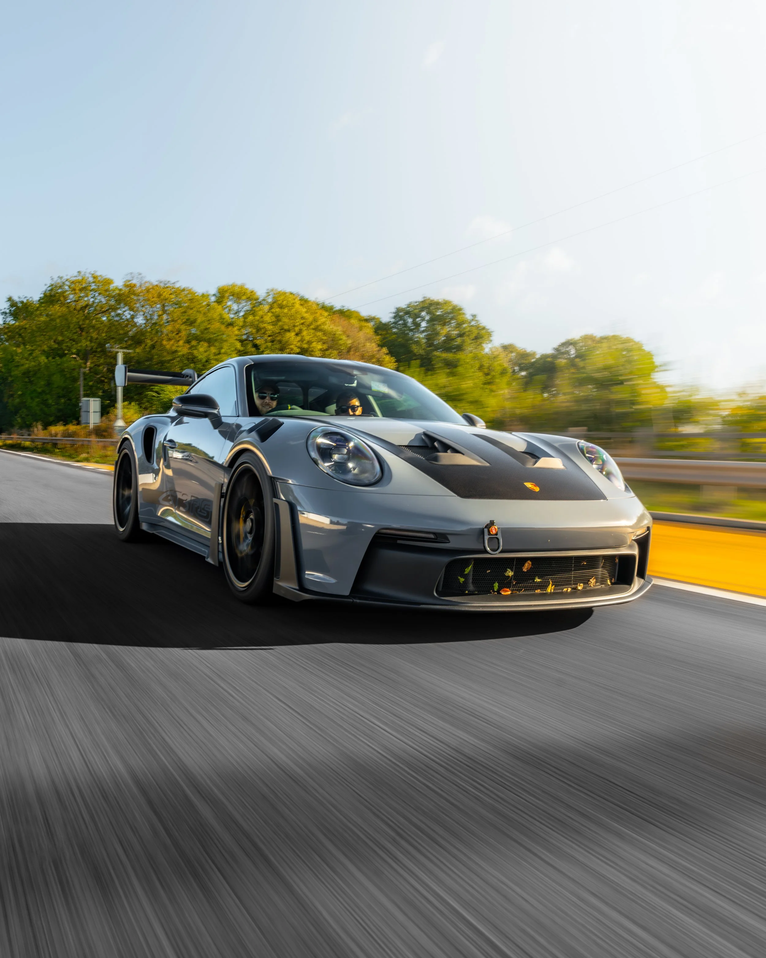 A silver Porsche sports car driving fast on a highway during daytime, with a large rear spoiler, black accents, and three people inside, trees and blue sky in the background.