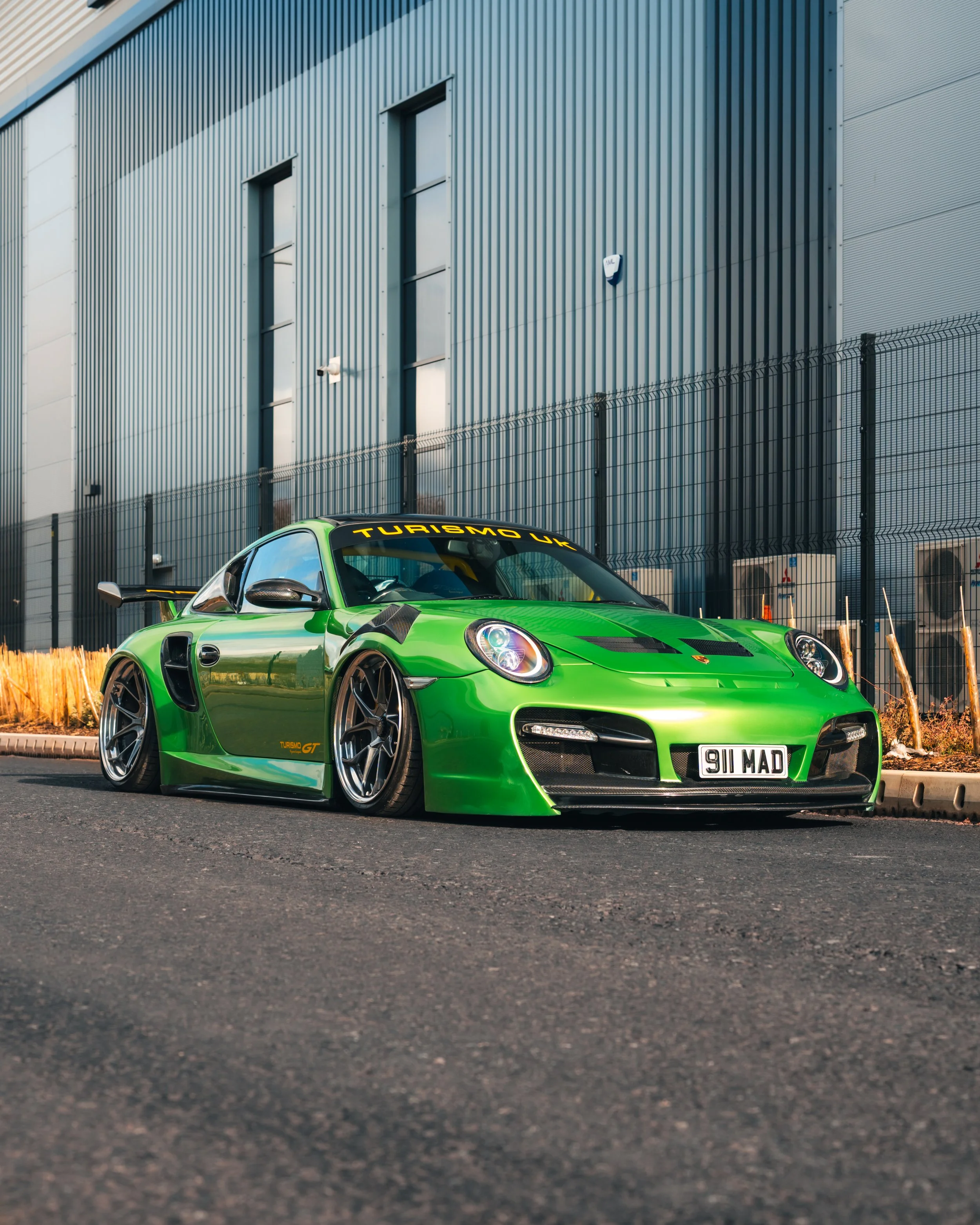 A bright green Porsche sports car with racing modifications parked on the side of the road in front of a modern industrial building with blue metal siding and a black metal fence.