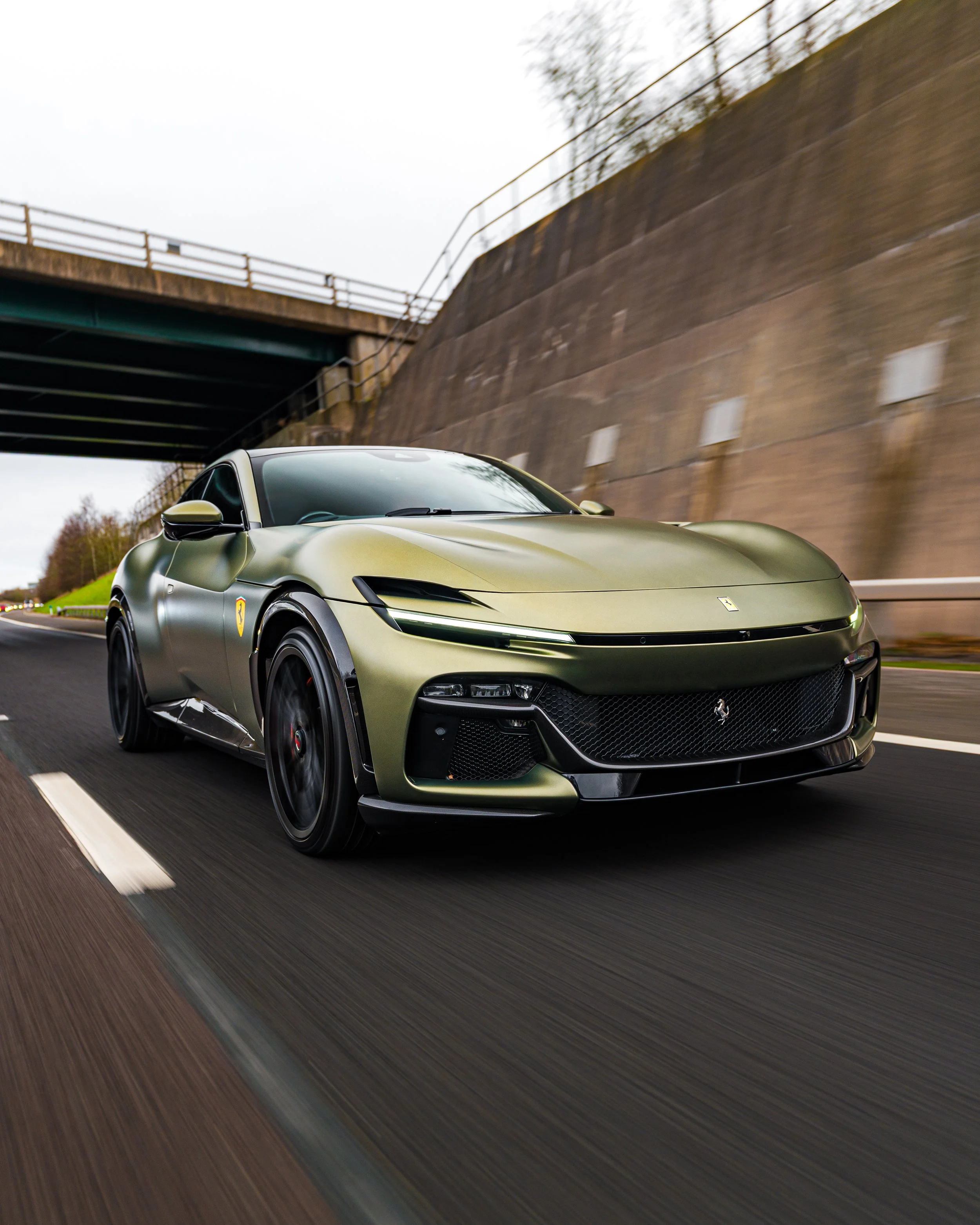 A sleek, matte green Ferrari sports car driving on a highway under an overpass.