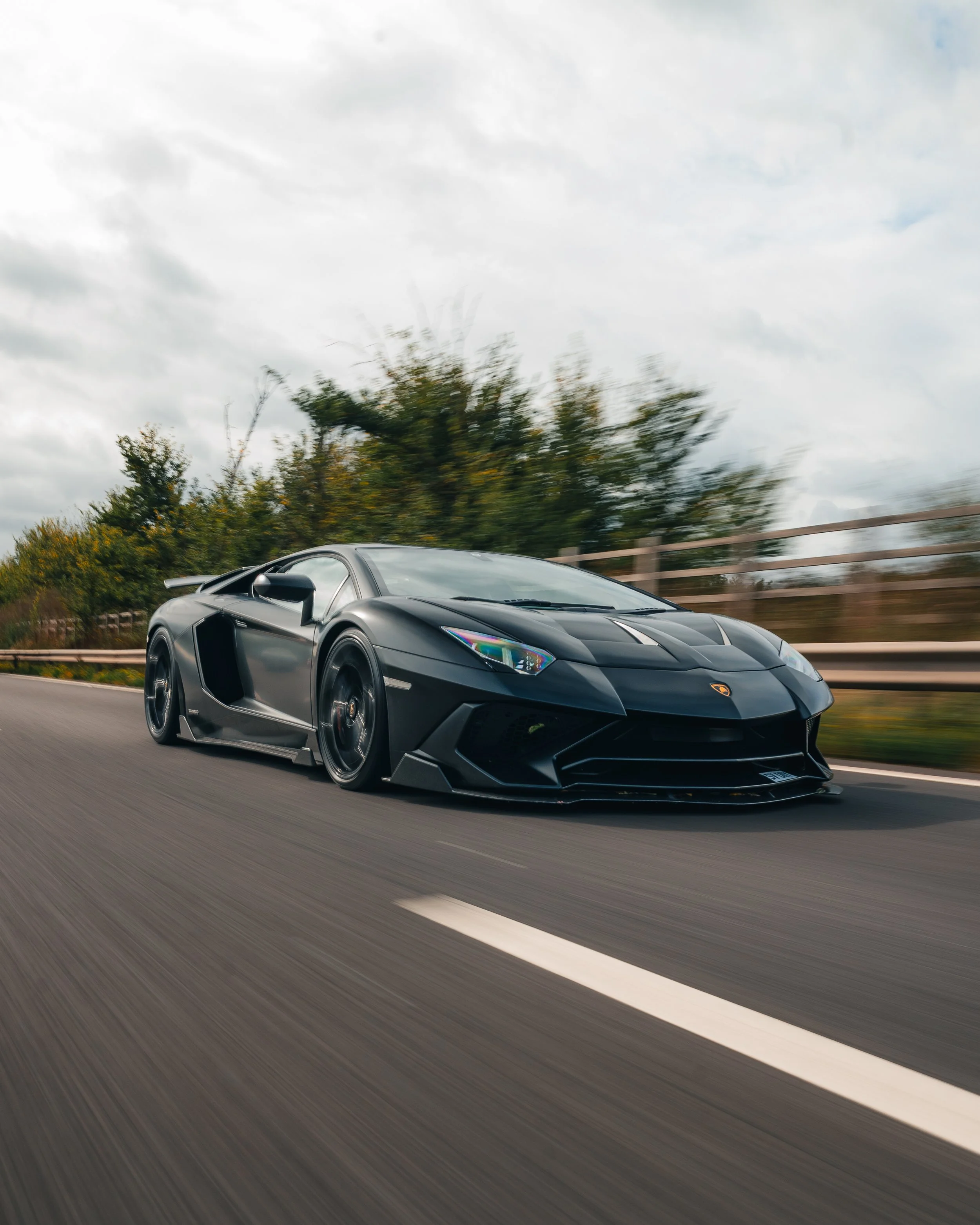 A black Lamborghini sports car racing on a highway with trees and cloudy sky in the background.