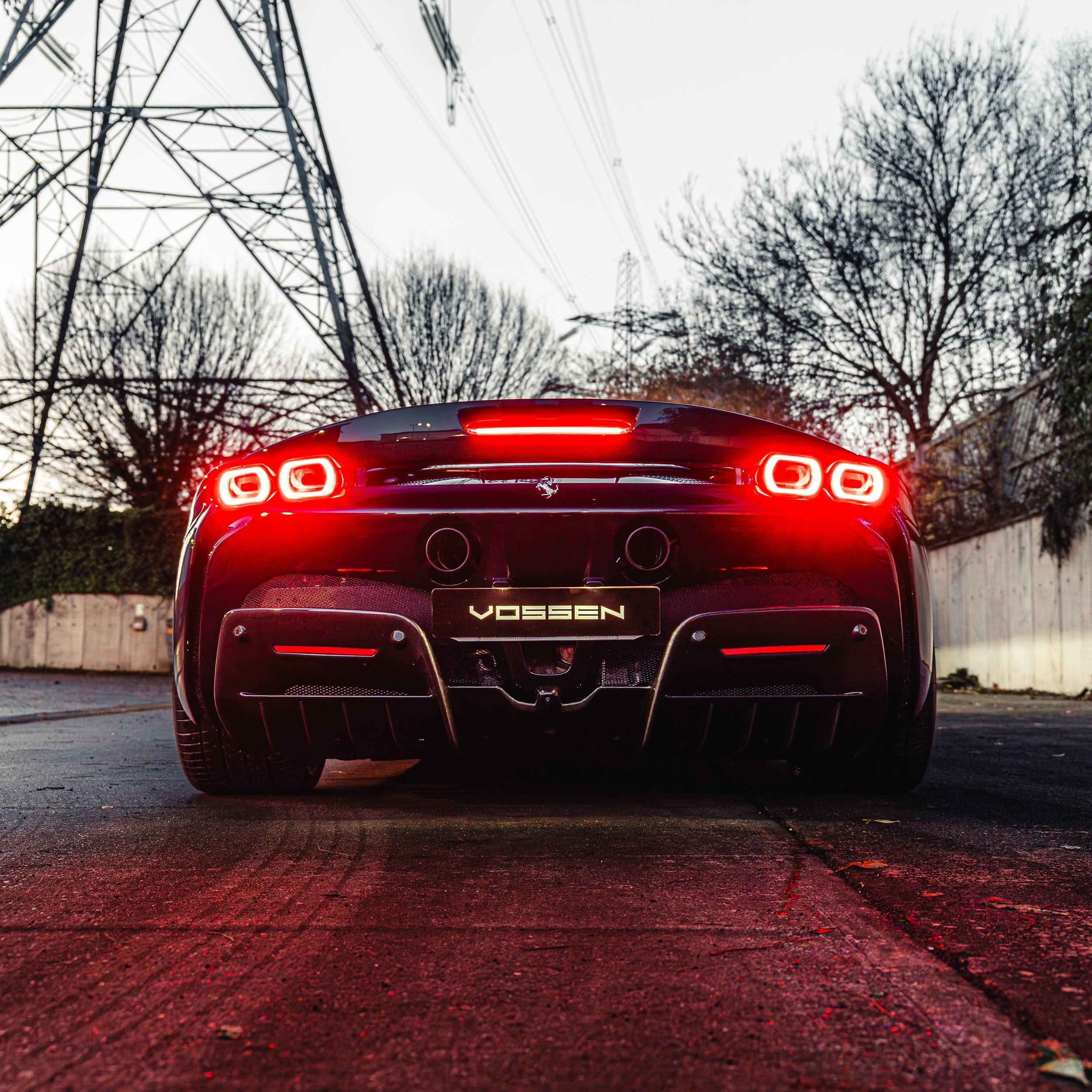 Rear view of a black Ferrari Vossen sports car with illuminated tail lights, parked on a street with overcast sky and leafless trees in the background.