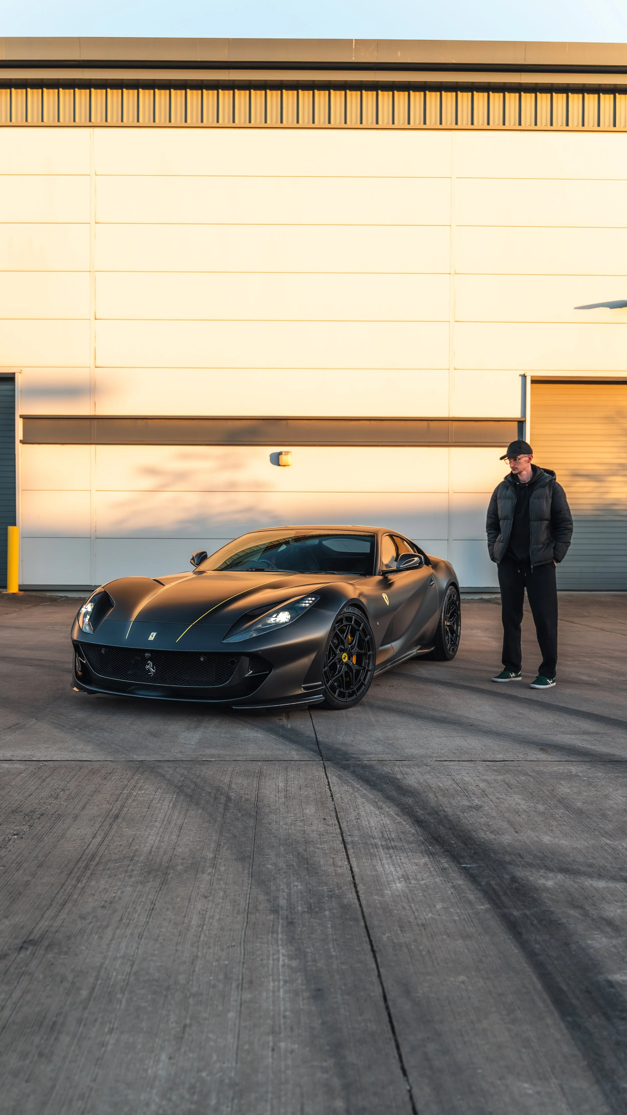 A man standing next to a matte black Ferrari sports car parked in front of a large industrial building during sunset.