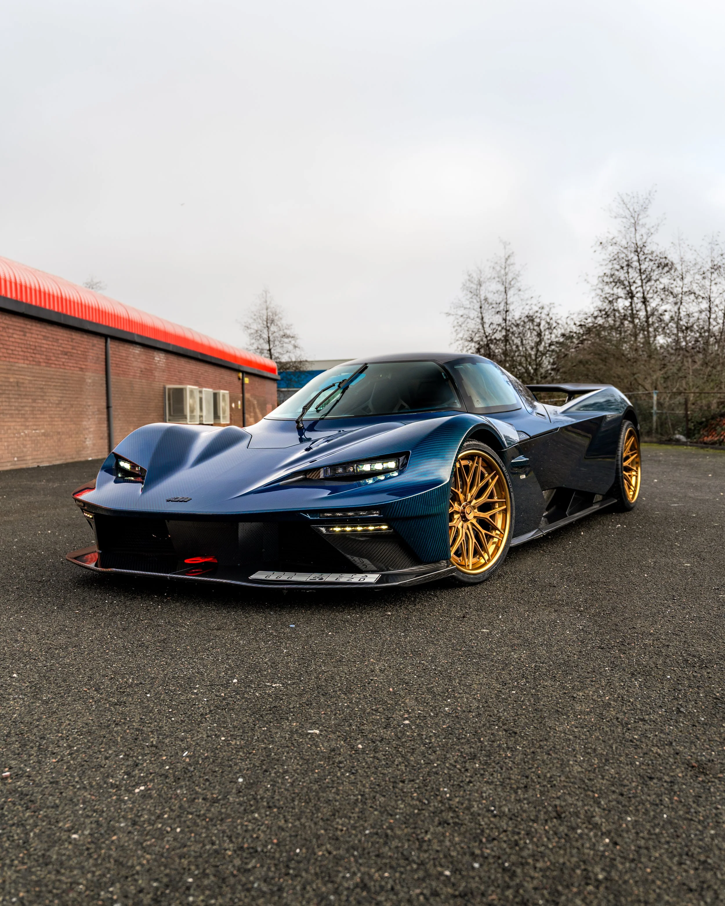 A sleek blue hypercar with gold wheels parked on asphalt, with a brick building with a red roof and air conditioning units, and leafless trees in the background.
