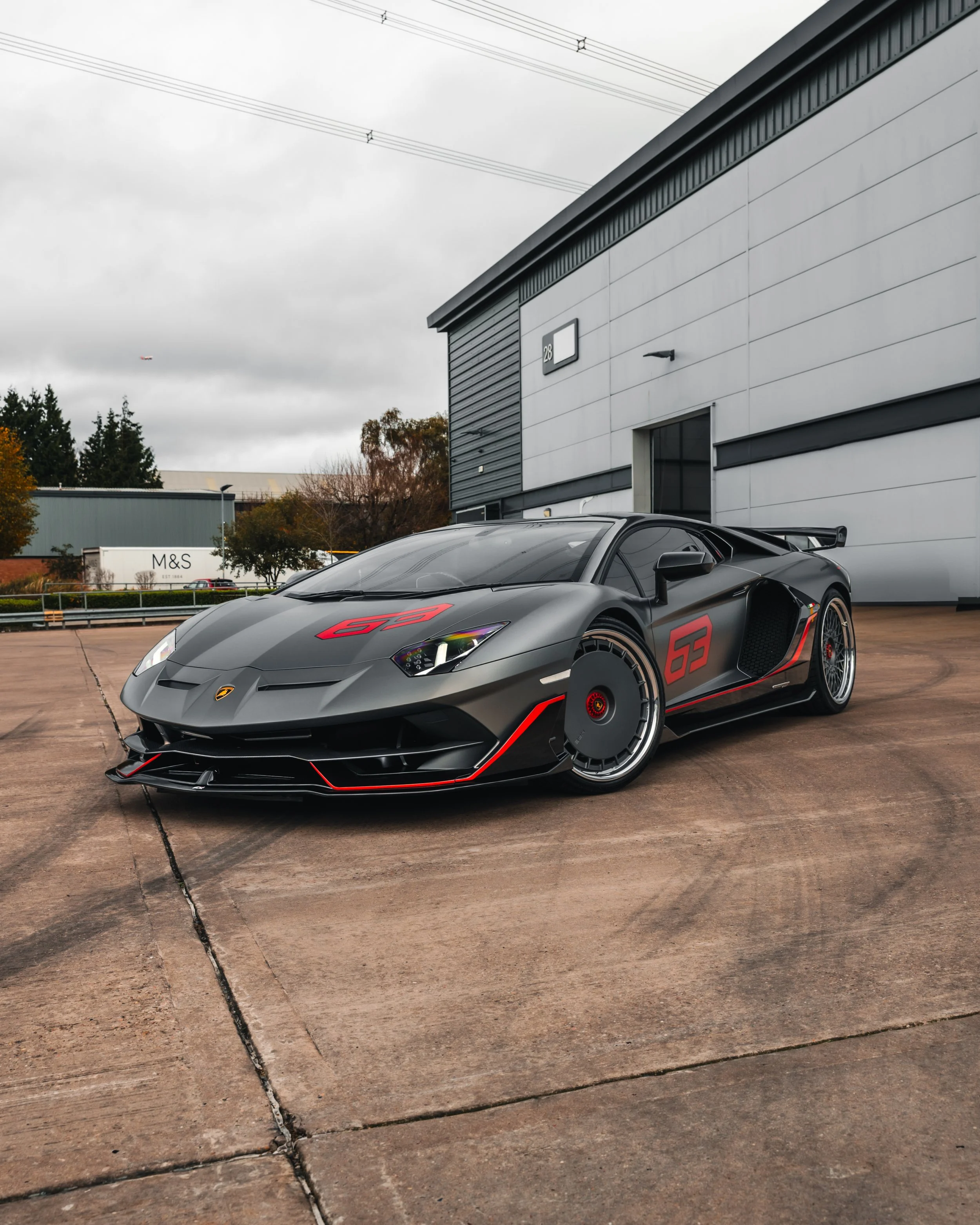 A matte black Lamborghini sports car with red accents and racing decals parked outdoors near a modern industrial building on a cloudy day.