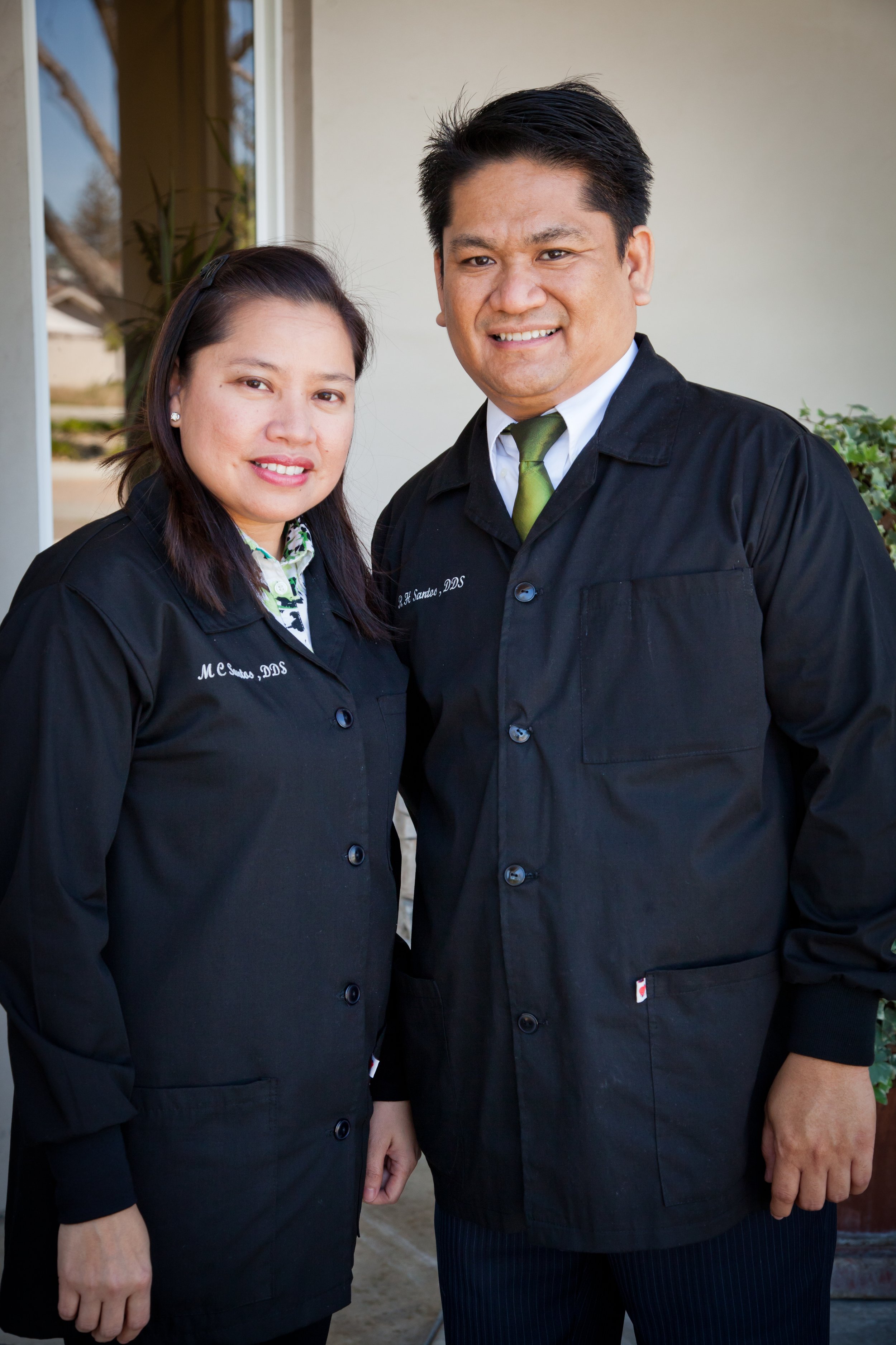 A man and a woman dressed in black chef coats standing outdoors in front of a beige wall. The man has dark hair and is wearing a tie; the woman has dark hair pulled back.