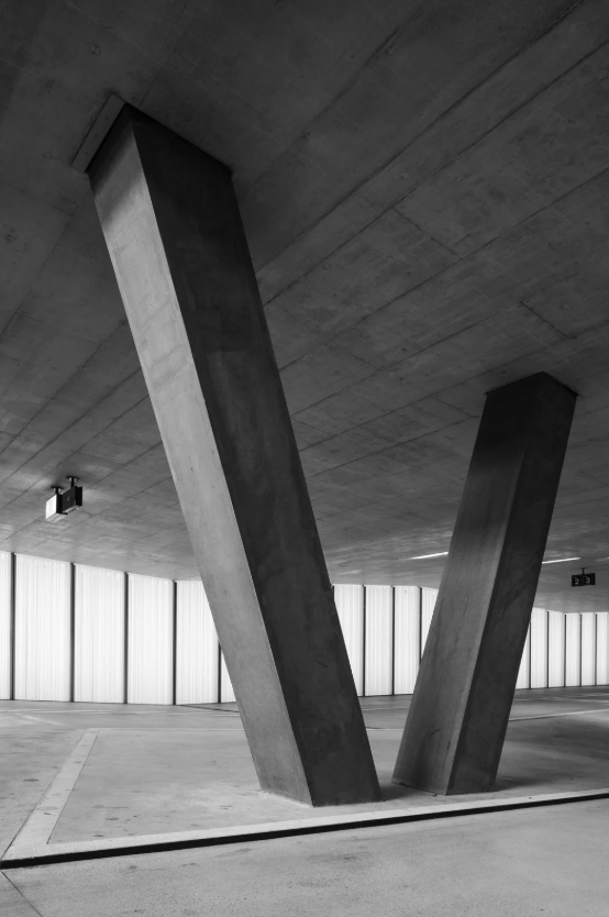 Modern architectural structure with two large angled concrete supports in an open parking area under a concrete ceiling.