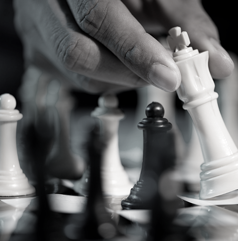 Close-up of a hand moving a white chess queen piece on a chessboard, with other black and white chess pieces visible.