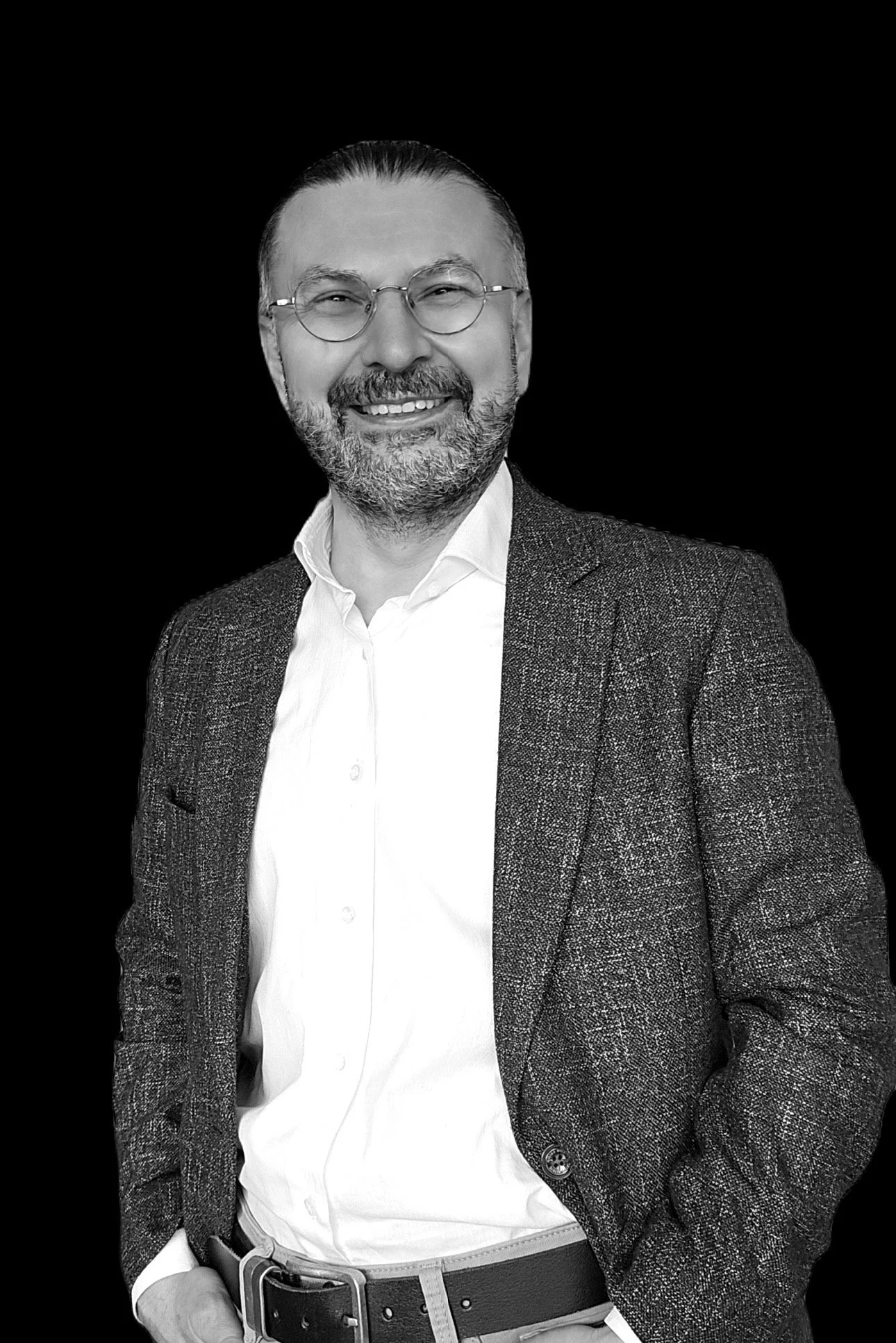 A smiling man with glasses, beard, and mustache, wearing a white shirt, blazer, and belt, posed against a black background.