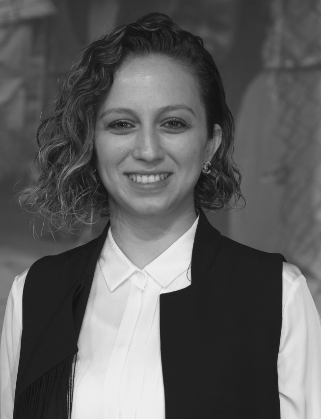 A woman with short, curly hair smiling at the camera, wearing a white collared shirt and a black vest, in a professional setting.