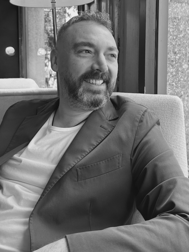 A smiling man with a beard and short hair, wearing a blazer and a white t-shirt, sitting on a cushioned seat near a window in a cafe or restaurant.