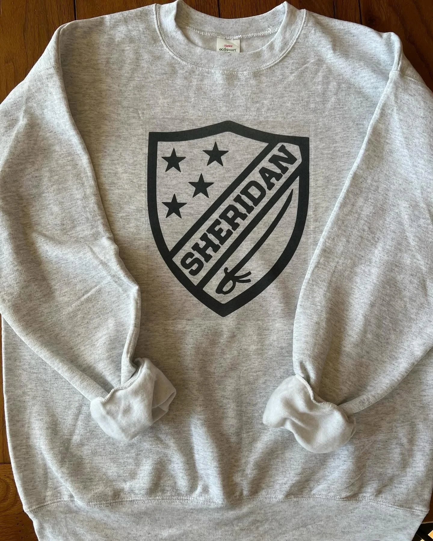 A light gray sweatshirt with a black shield logo featuring five stars and the word SHERIDAN across a diagonal stripe.