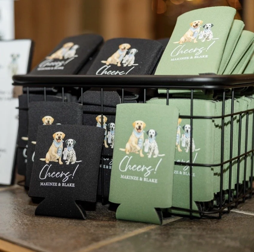 A display of drink koozies with illustrations of two dogs and the text "Cheers! MAKINZE & BLAKE" on a metal rack.
