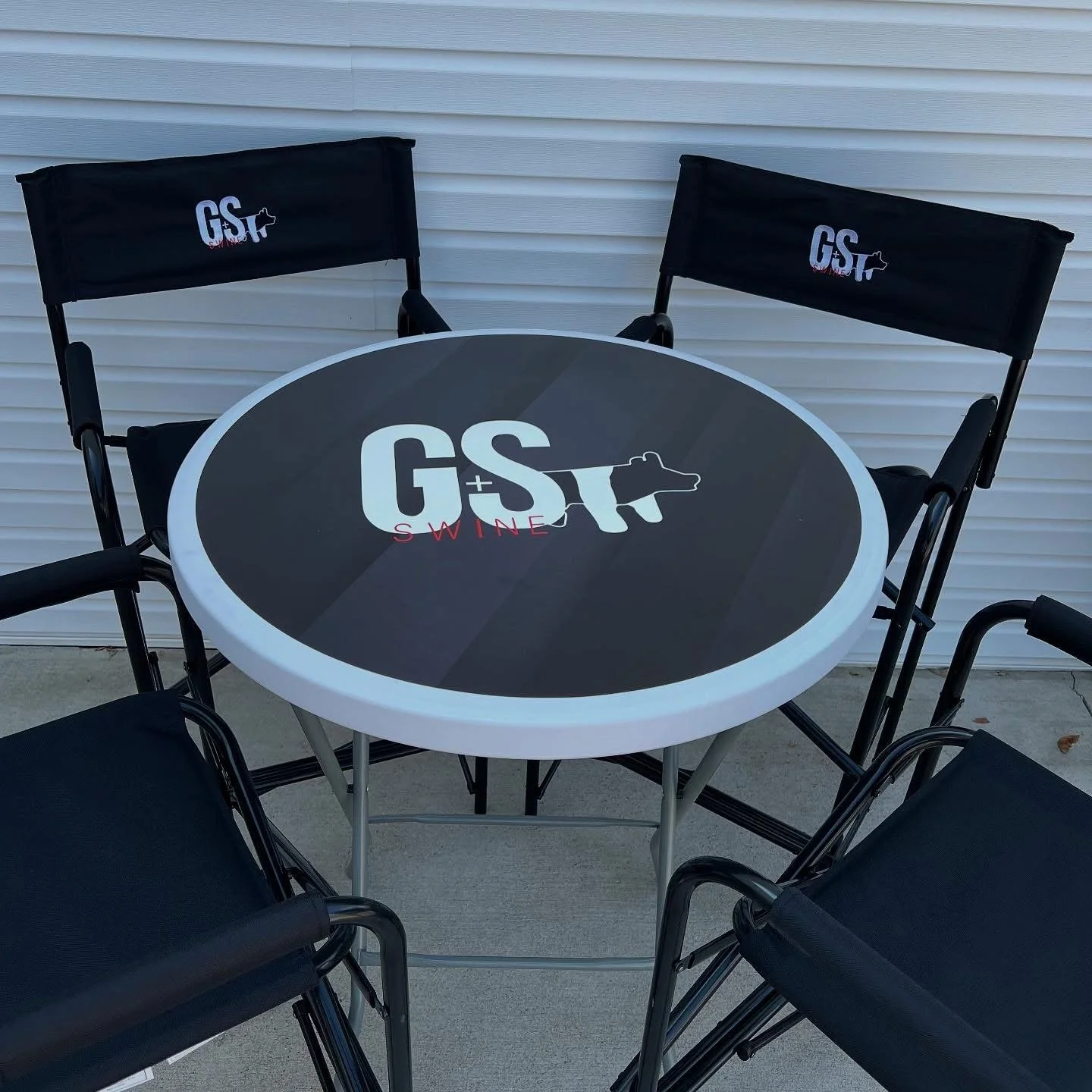 Outdoor round table with a black top and white border, featuring the 'G+S SWINE' logo, surrounded by four black chairs, in front of a white garage door.
