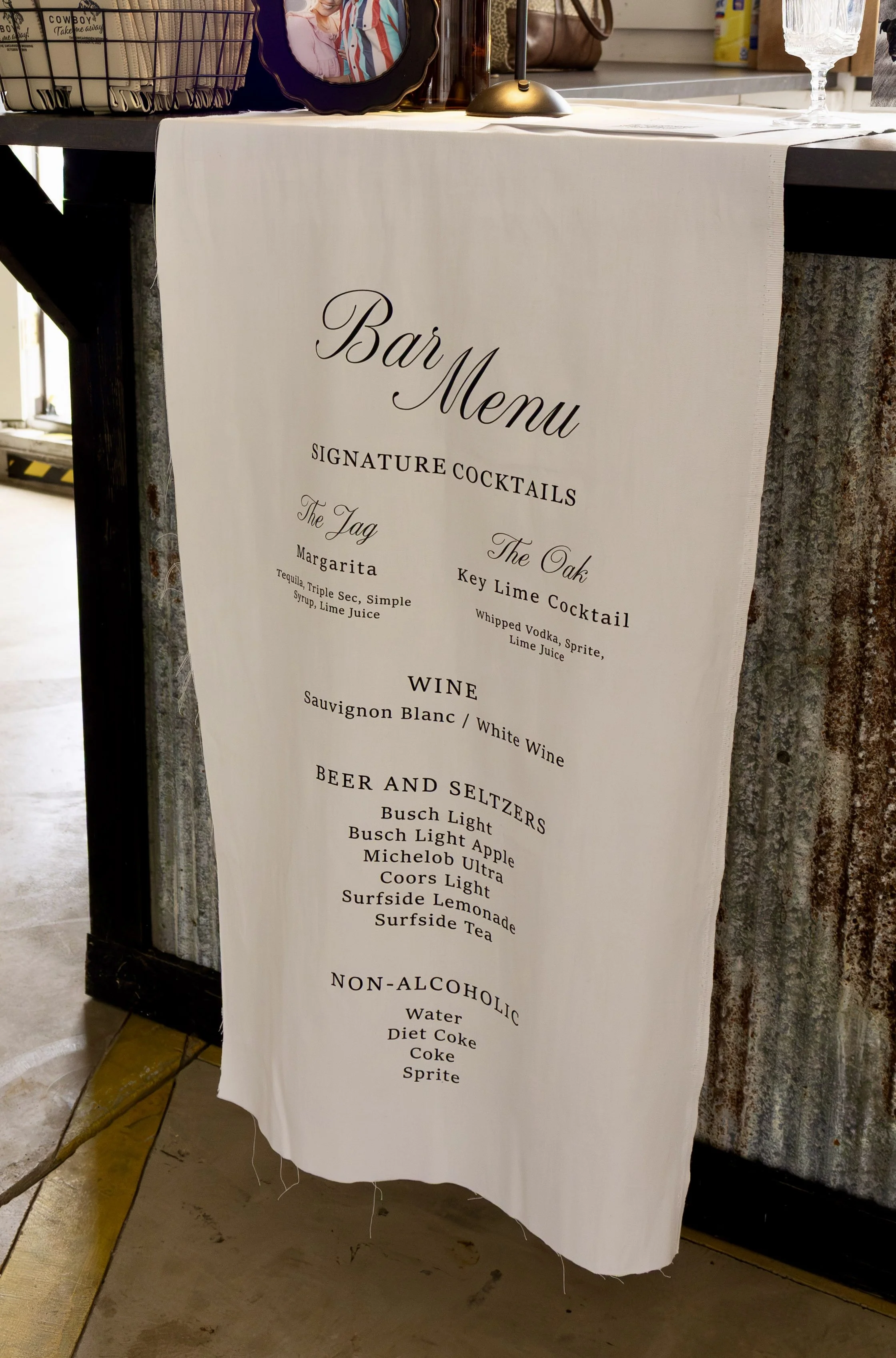 A white fabric banner displaying a bar menu with signature cocktails, wines, beers, and non-alcoholic drinks, hung on a wooden stand at a bar or restaurant.