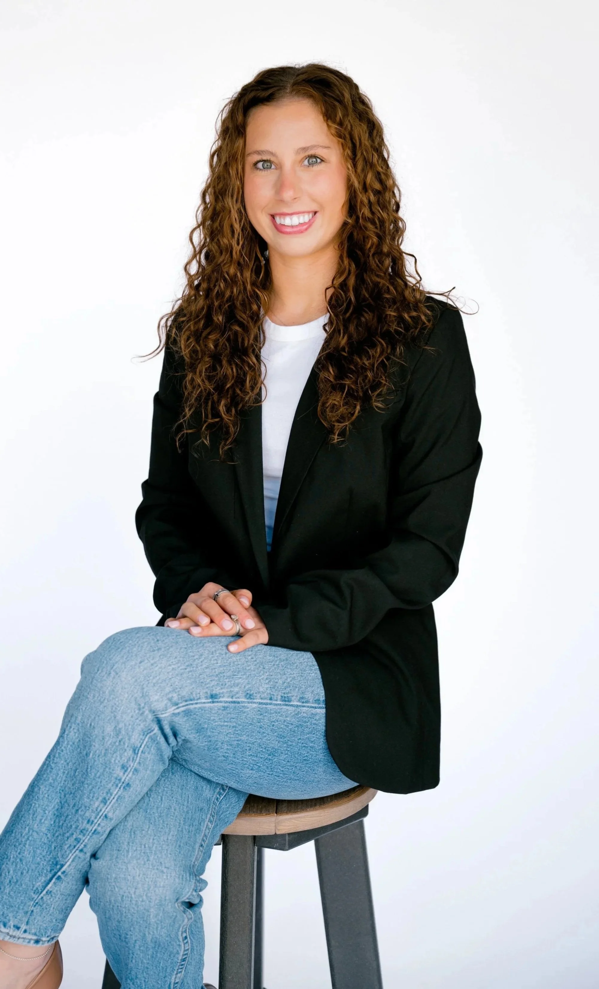 A woman with curly brown hair sitting on a wooden stool against a white background, smiling, wearing a black blazer, white shirt, and light blue jeans.