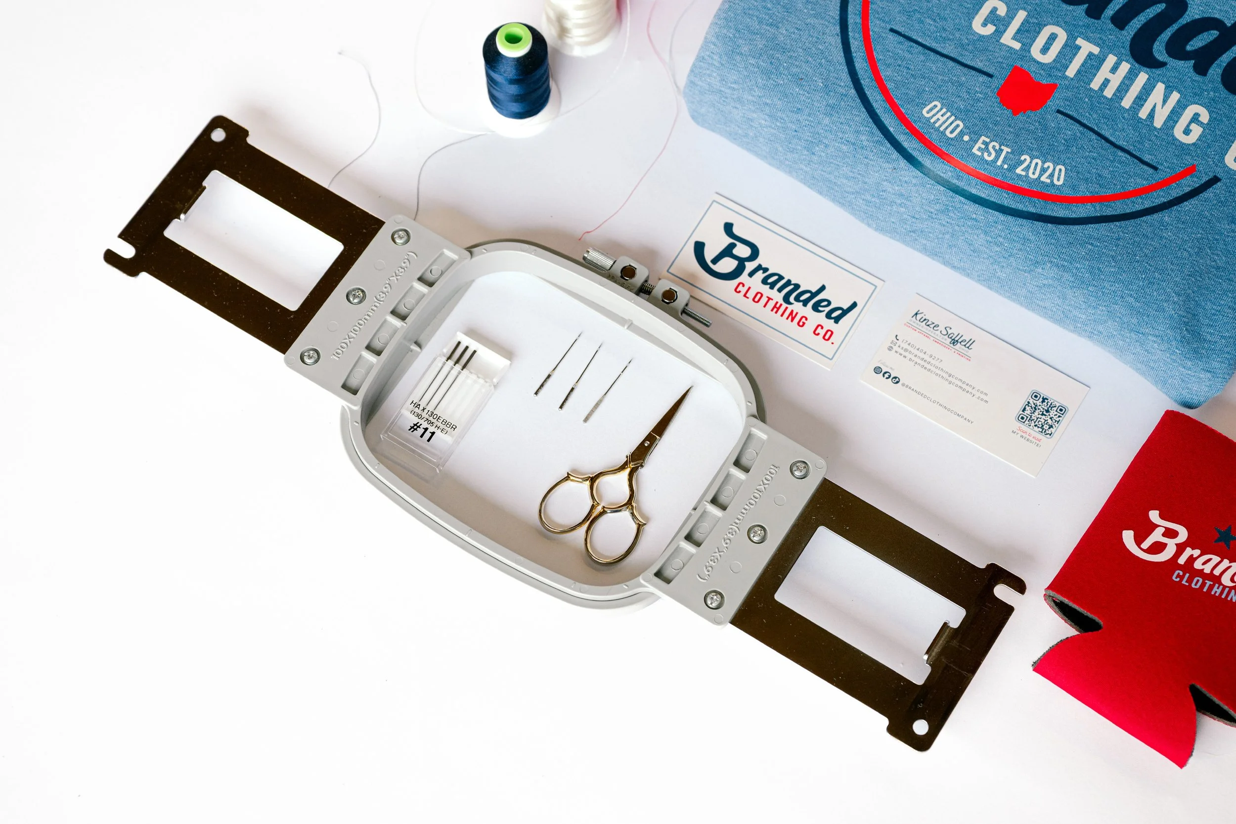 Sewing machine frame with sewing scissors, needle puller, and sewing threads beside a blue clothing box with 'Granded Clothing Co.' logo and a red sock partially visible in the bottom right corner.