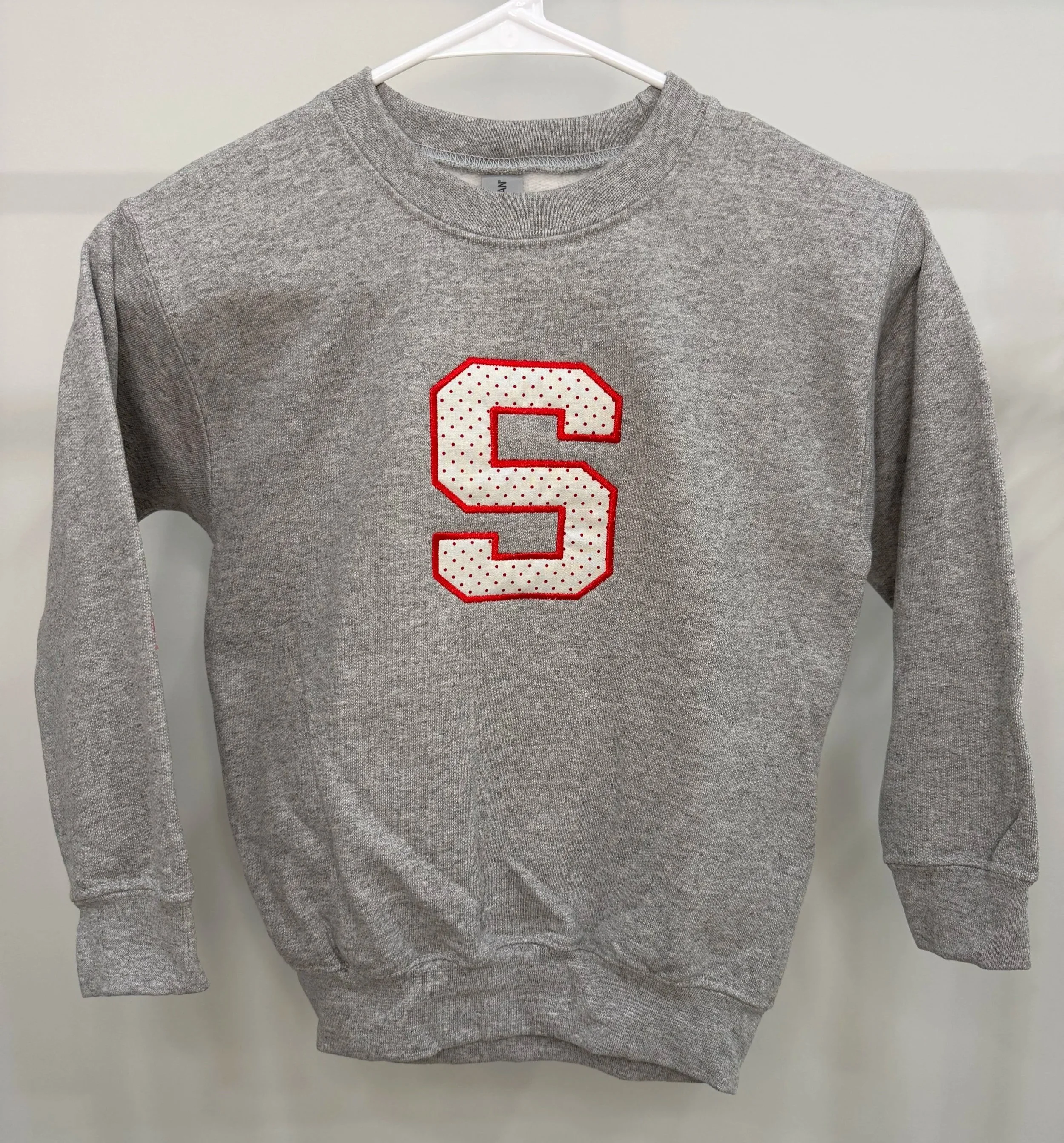Gray sweatshirt with a red and white Cleveland State logo, hanging on a white hanger against a beige wall.