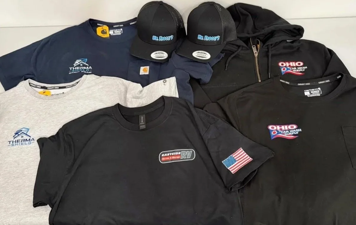 Various apparel and hats, including T-shirts with logos, a black hoodie, and black caps with blue text, displayed on a flat surface.