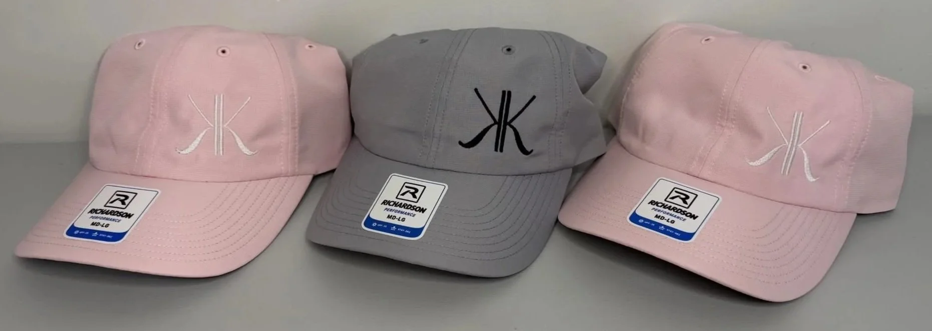 Three baseball caps, two pink with white logo and one gray with black logo, displayed on a surface.