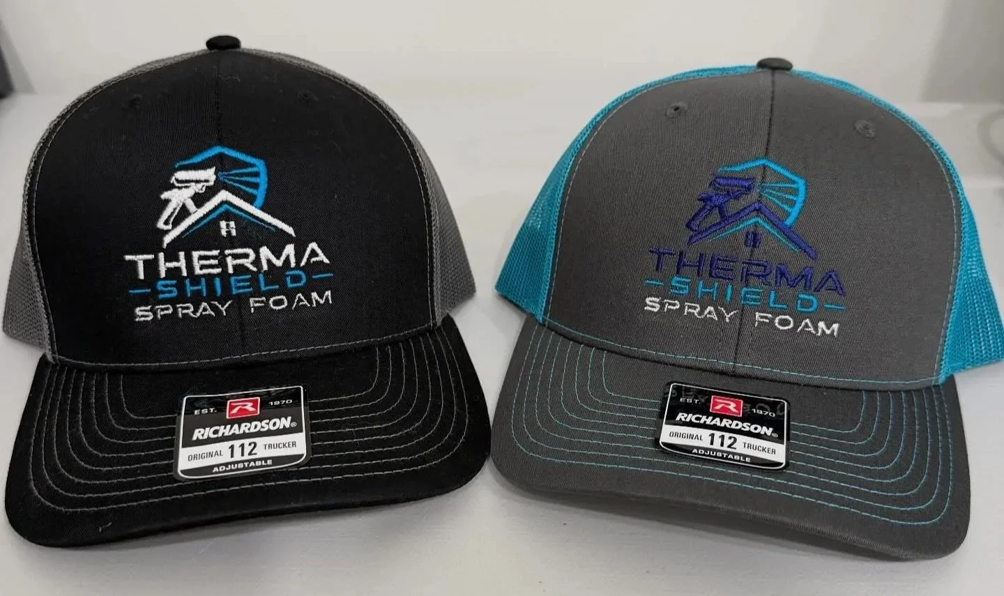 Two Richardson 112 trucker hats with embroidered logos and text, one black with gray mesh and one gray with blue mesh, on a white surface.