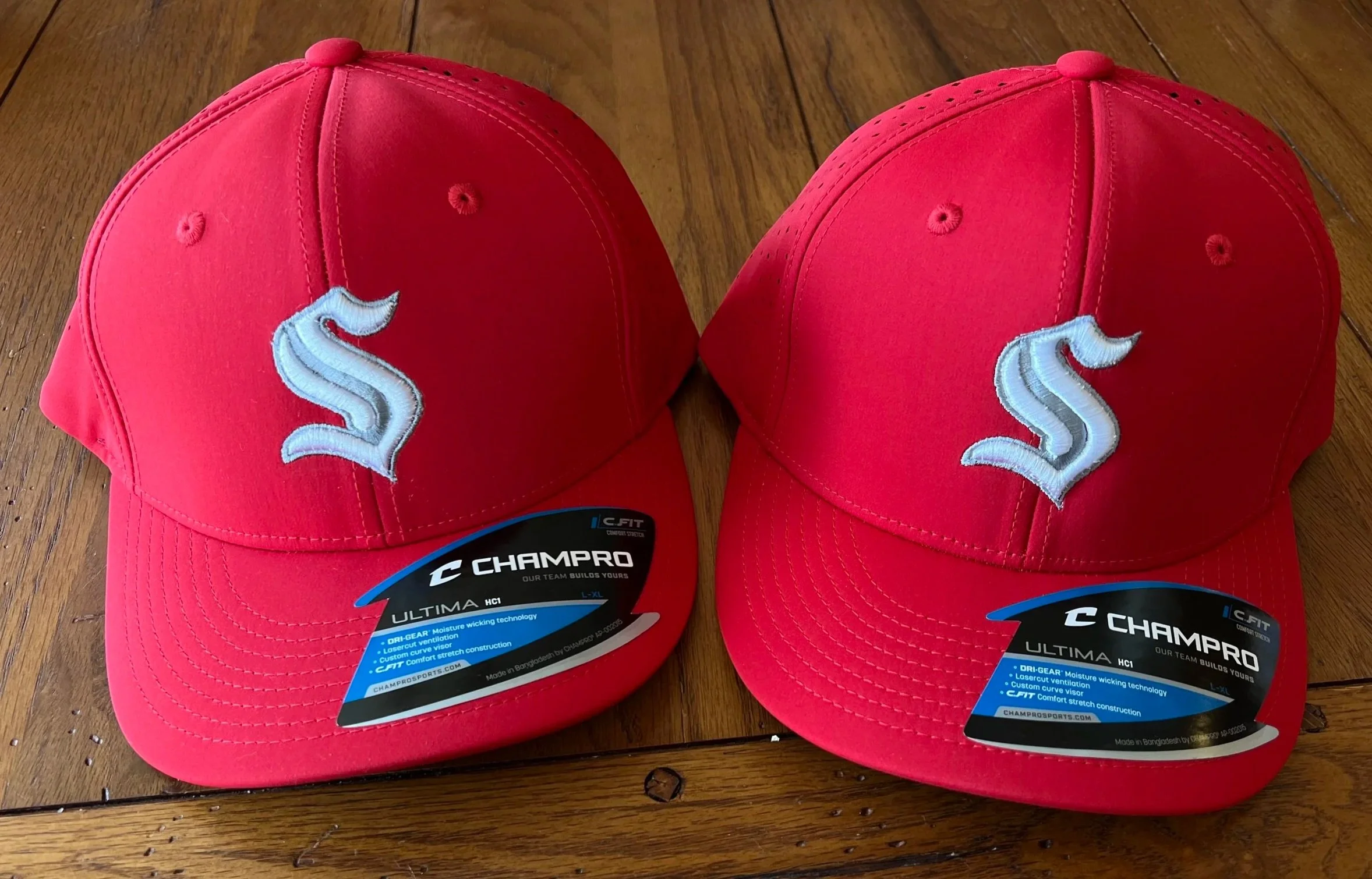 Two red baseball caps with a white embroidered logo on the front, placed on a wooden surface. The caps have tags and labels attached, indicating they are from the brand CHAMPRO.