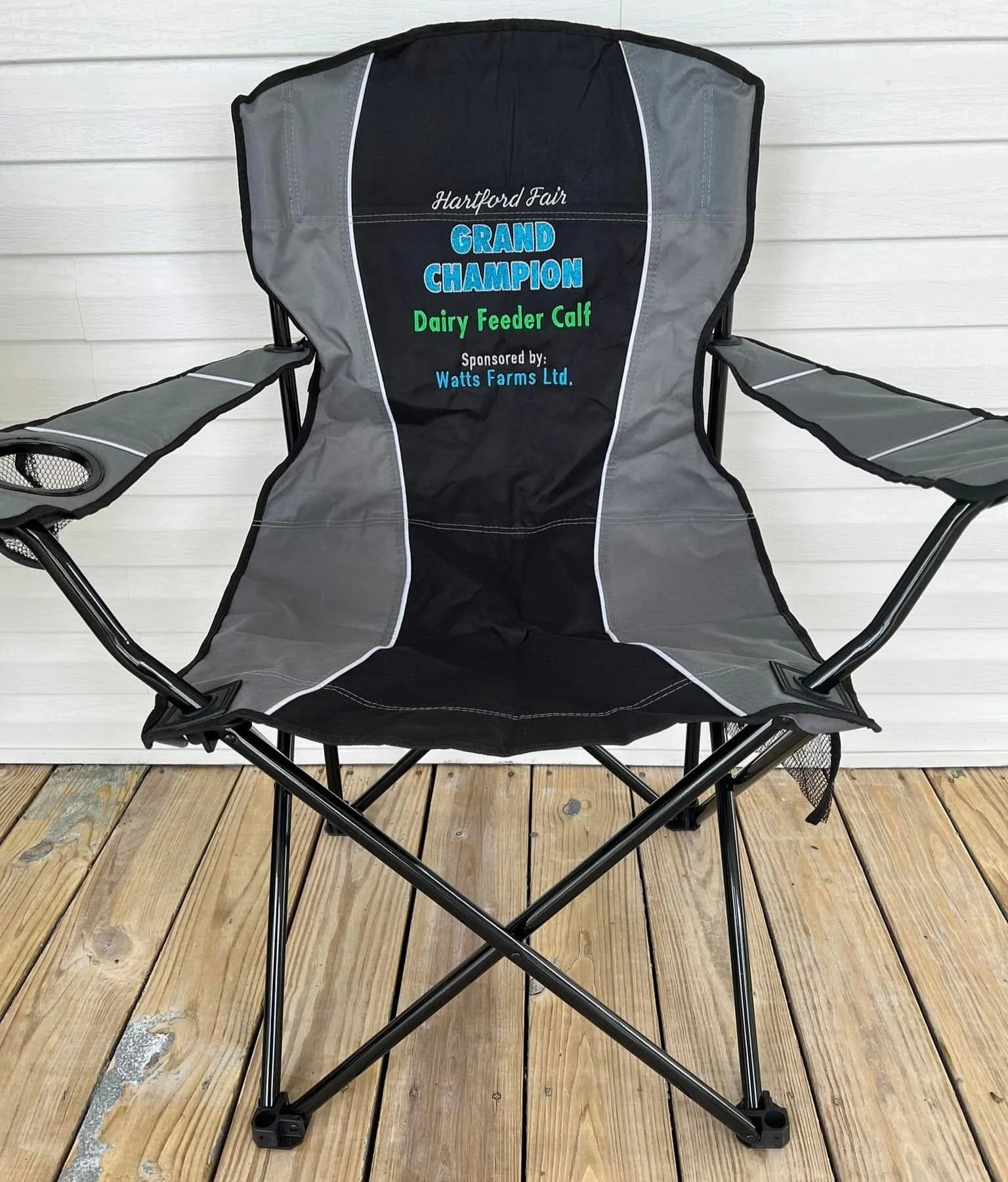 Portable folding chair with a black and gray fabric, featuring embroidered text about a dairy feeder calf and sponsor on the backrest, placed on a wooden deck against a white wall.