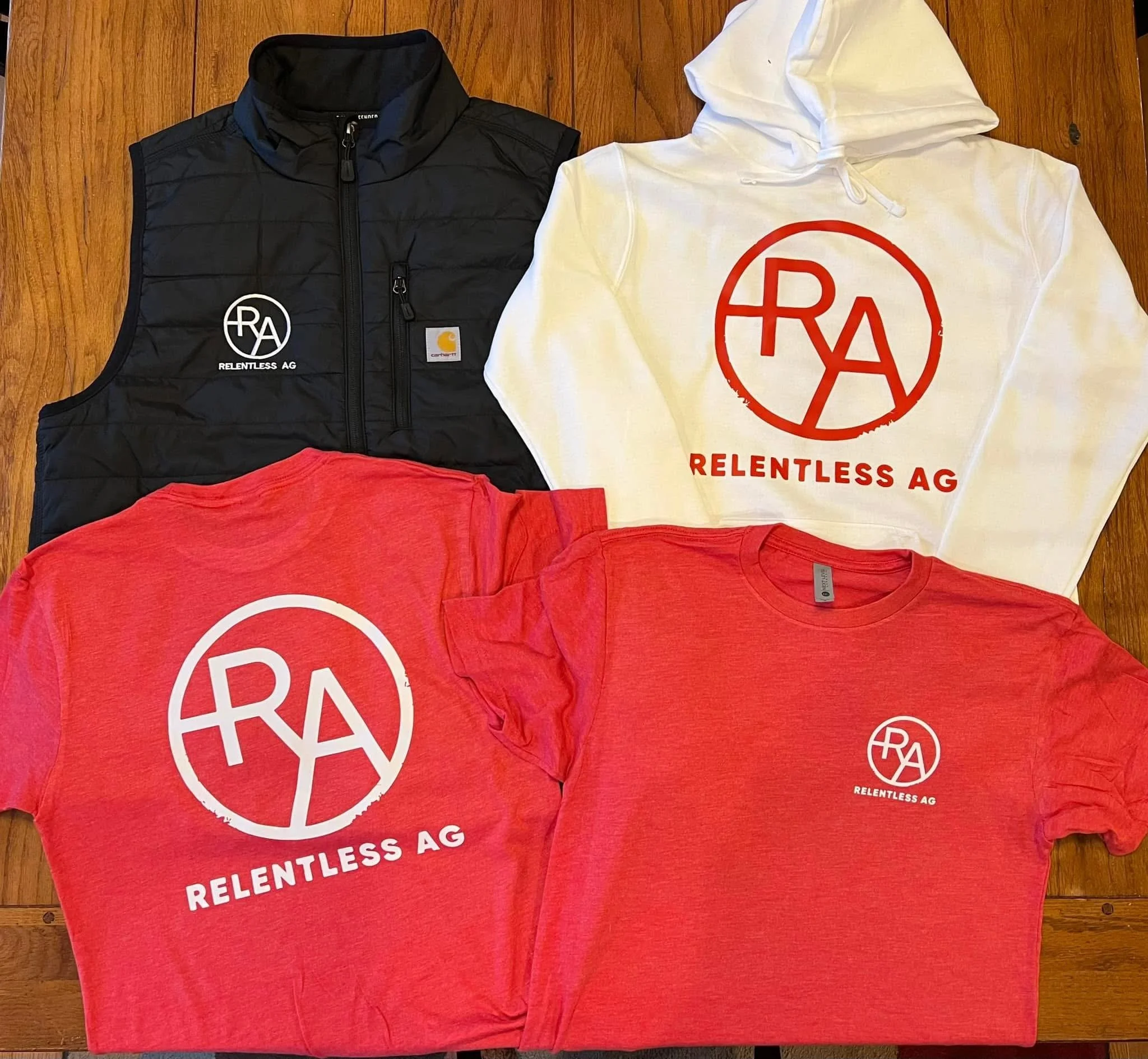 A black vest and a white hoodie, both with the Red Bull Racing Alliance logo and the words "Relentless AG," are lying on a wooden surface, with a red T-shirt partially visible in front of them.