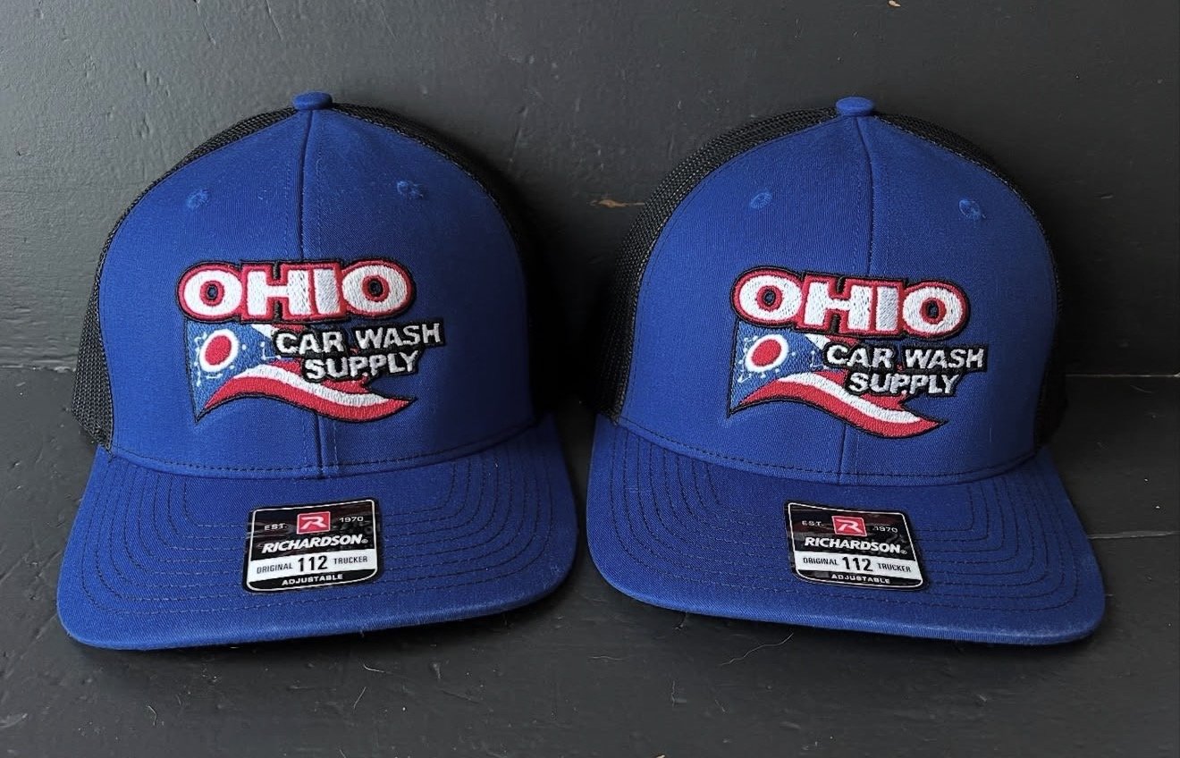 Two blue trucker caps with 'Ohio Car Wash Supply' embroidery and a stylized wave and bubble design. The caps are placed on a dark surface against a gray wall.