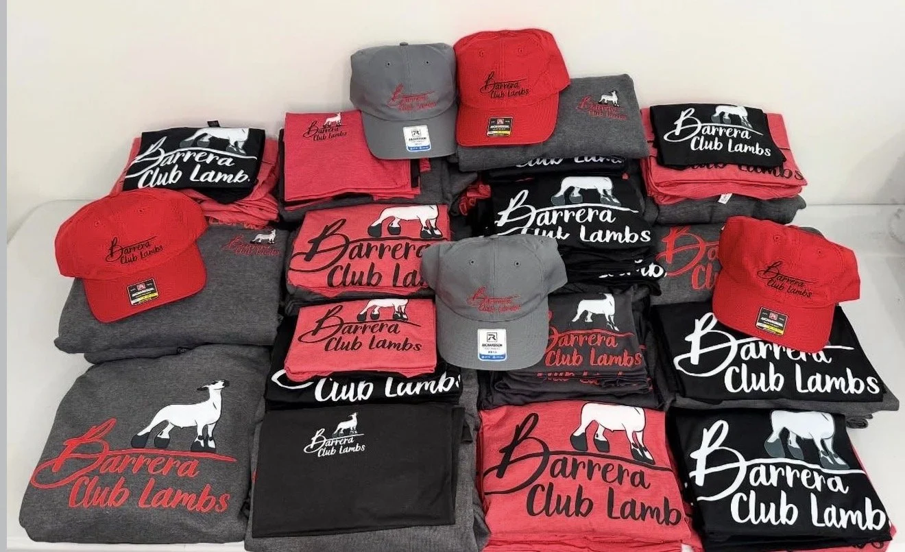 A display of clothing and hats with the logo 'Barrera Club Lambs,' including caps, t-shirts, and sweatshirts in red, black, and gray, arranged on a table.