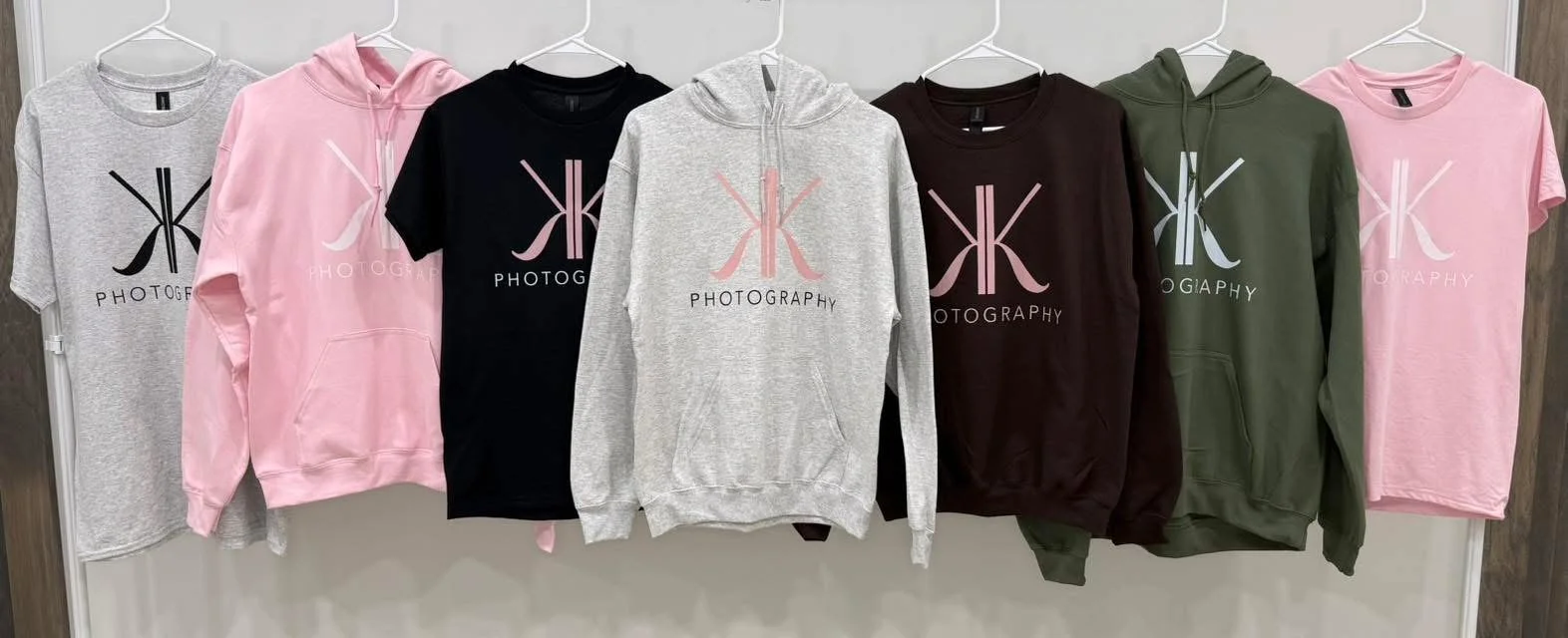 Six hoodies and t-shirts hanging on a white clothing rack. The clothing features a logo with the initials 'JK' and the word 'PHOTOGRAPHY'. The colors of the items are grey, pink, black, grey, green, and pink.